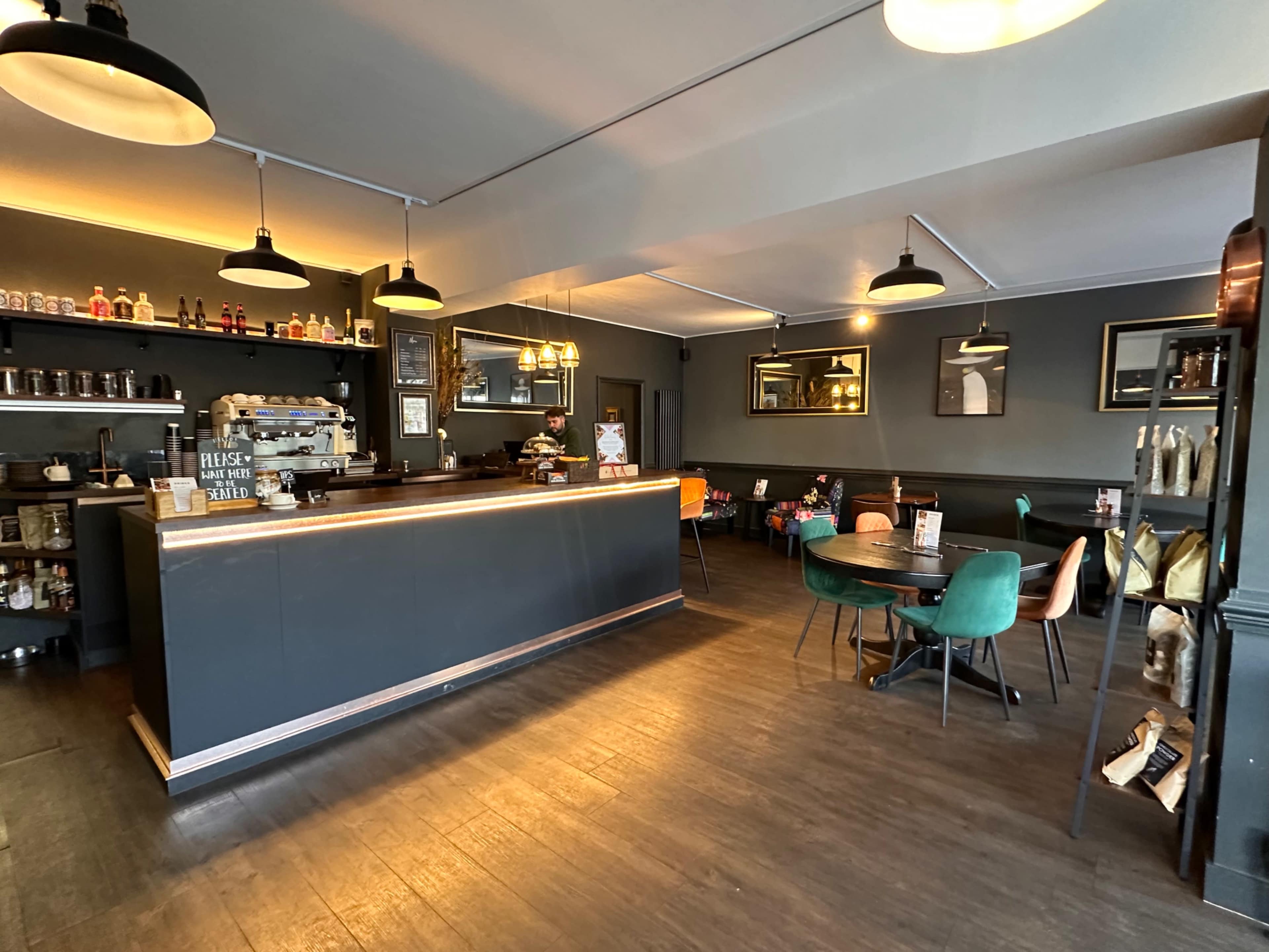 The image shows a modern café interior with a dark color scheme featuring a bar counter, seating areas, and shelves lined with bottles.
