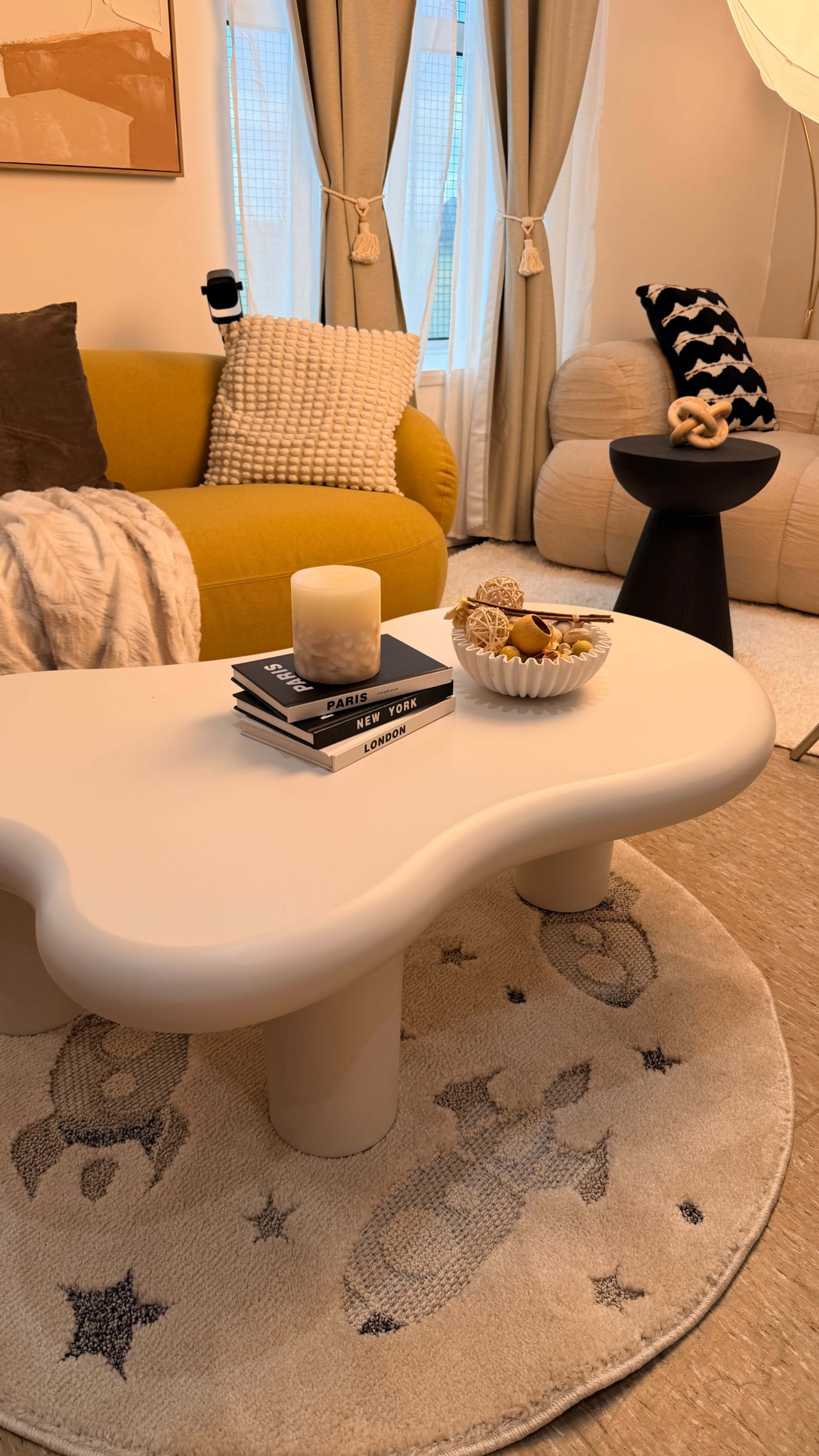 The image features a modern living room with a cloud-shaped coffee table, a yellow sofa, a black side table, and decorative elements including a bowl of snacks and stacked books.