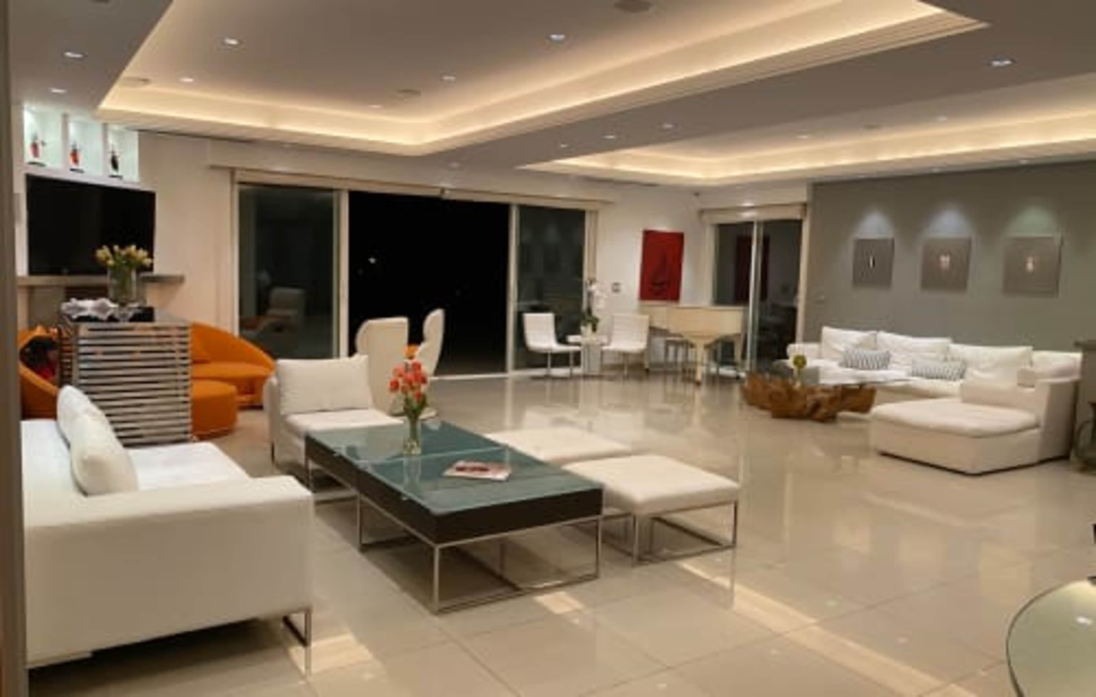 A modern, spacious living room with white furniture, a coffee table, and large windows that provide a view of the night sky.