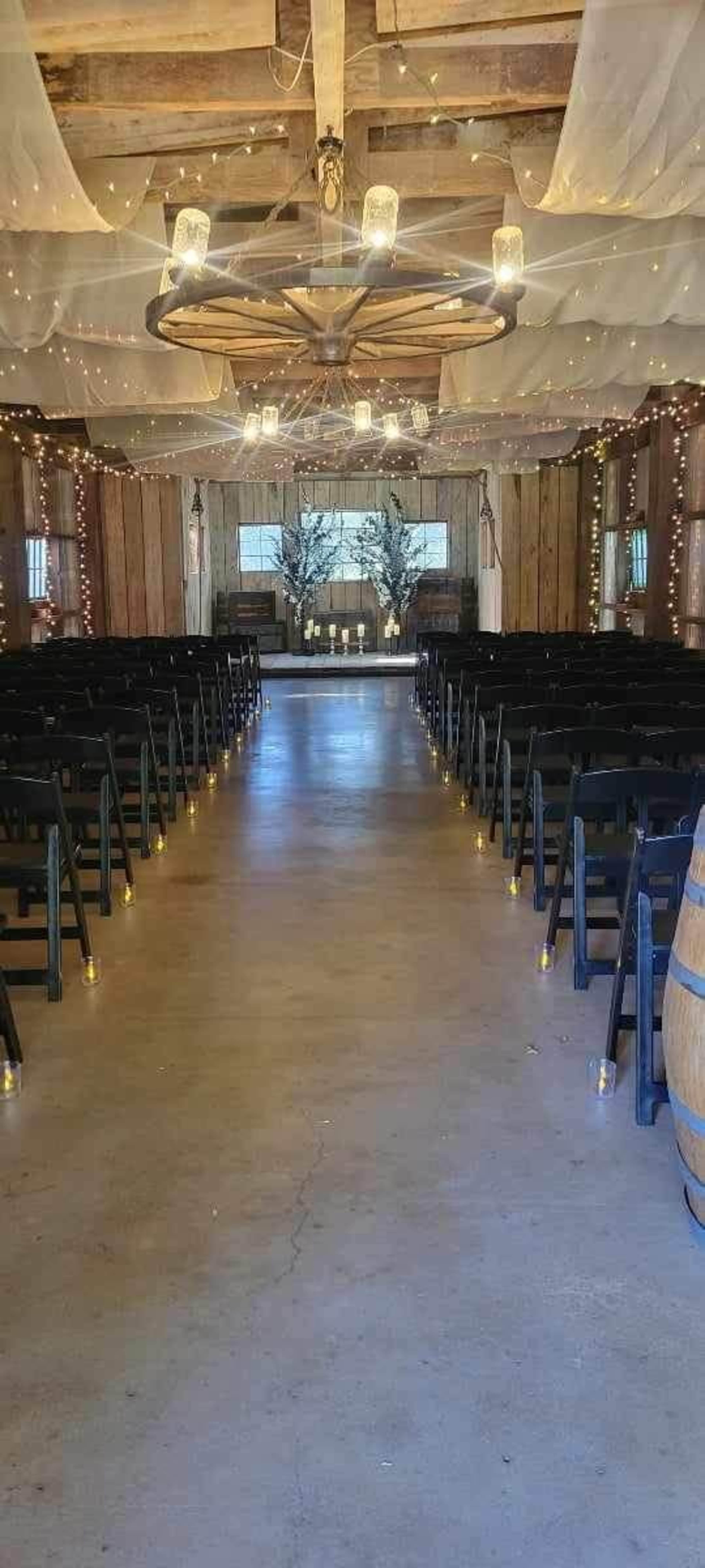 The interior of a rustic venue is set up for a wedding ceremony, featuring rows of black chairs, decorative lighting, and a decorated altar at the far end.