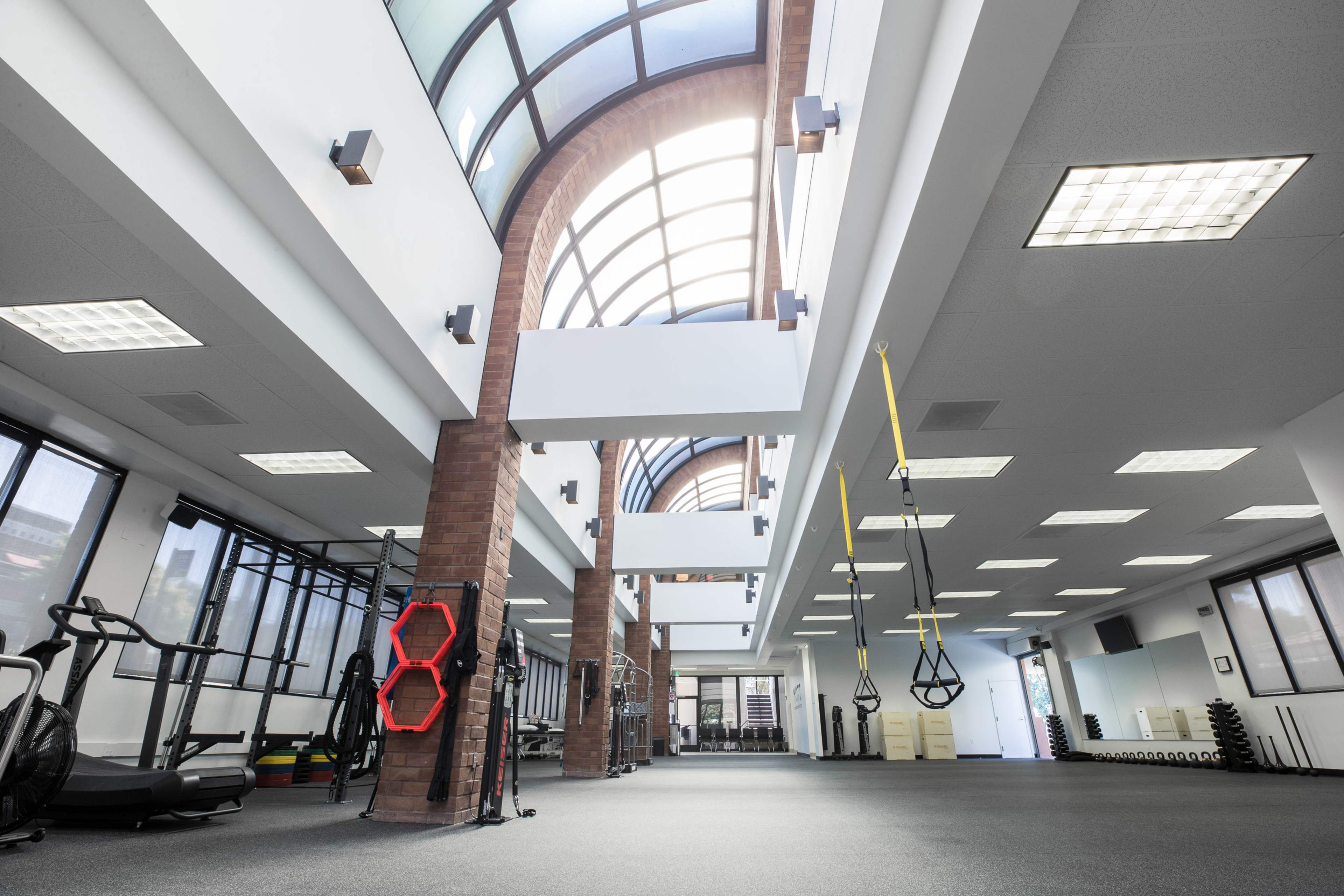 The image shows a spacious gym interior with large windows, exercise equipment, and suspended fitness straps.