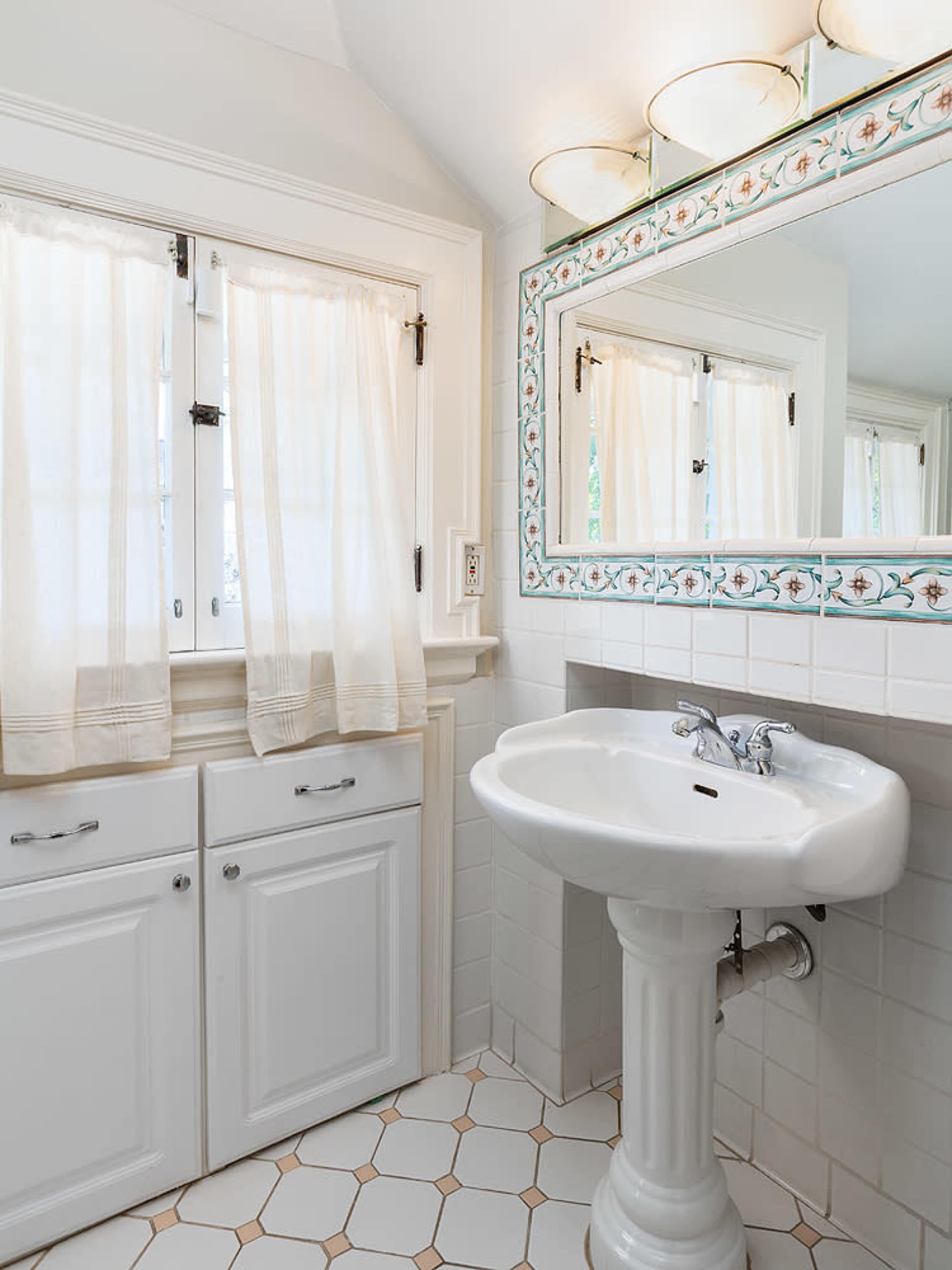 The image shows a bathroom with white cabinetry, a pedestal sink, a mirror above the sink, and light-colored curtains covering the windows.