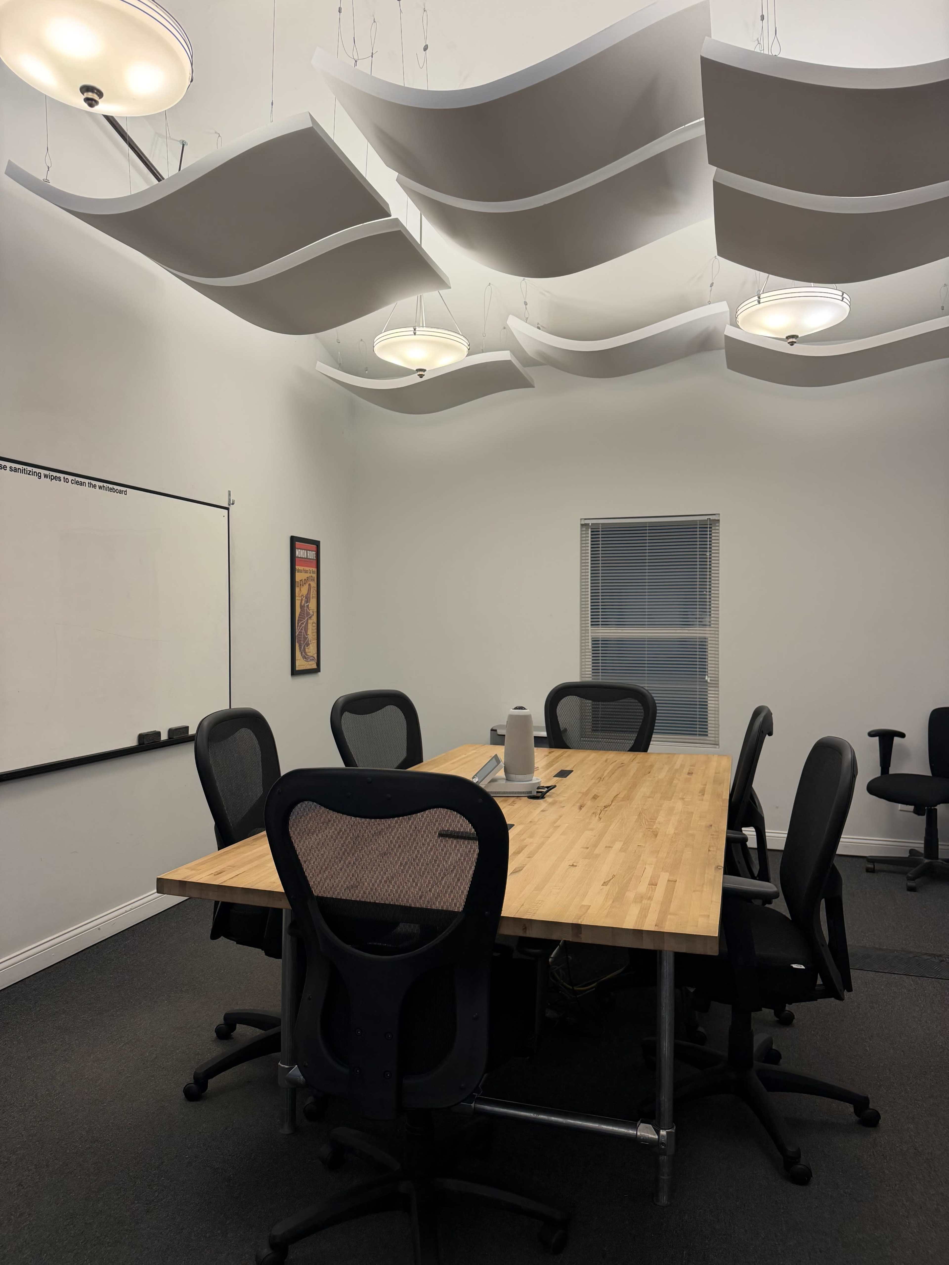 A conference room features a large wooden table surrounded by black office chairs, with white acoustic panels and light fixtures hanging from the ceiling.