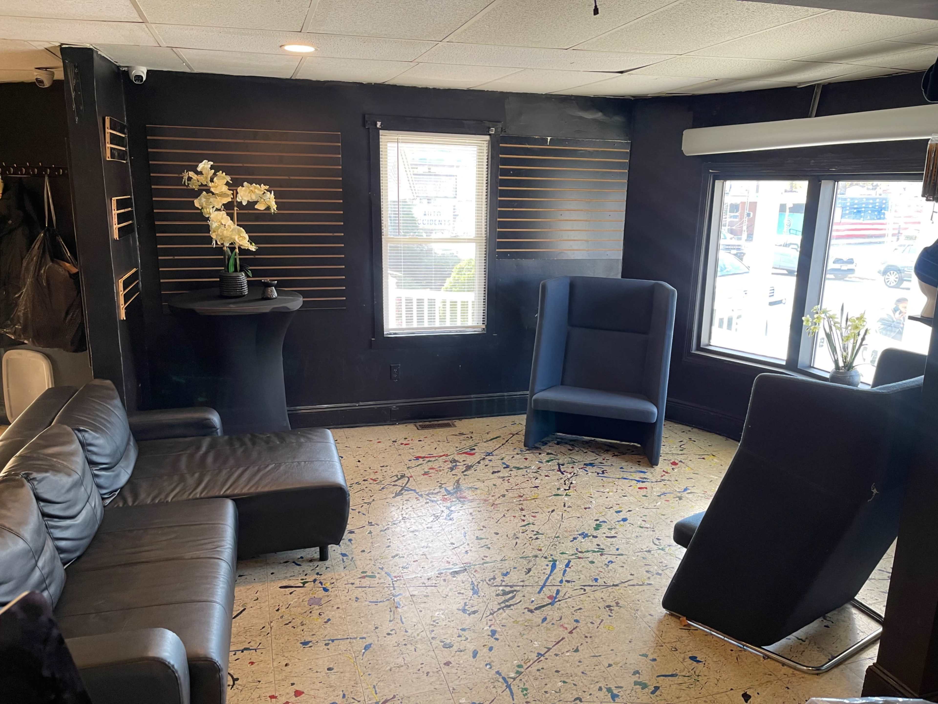 The image shows a sparsely furnished room with a black wall, a black leather sofa, and two blue chairs near a window.