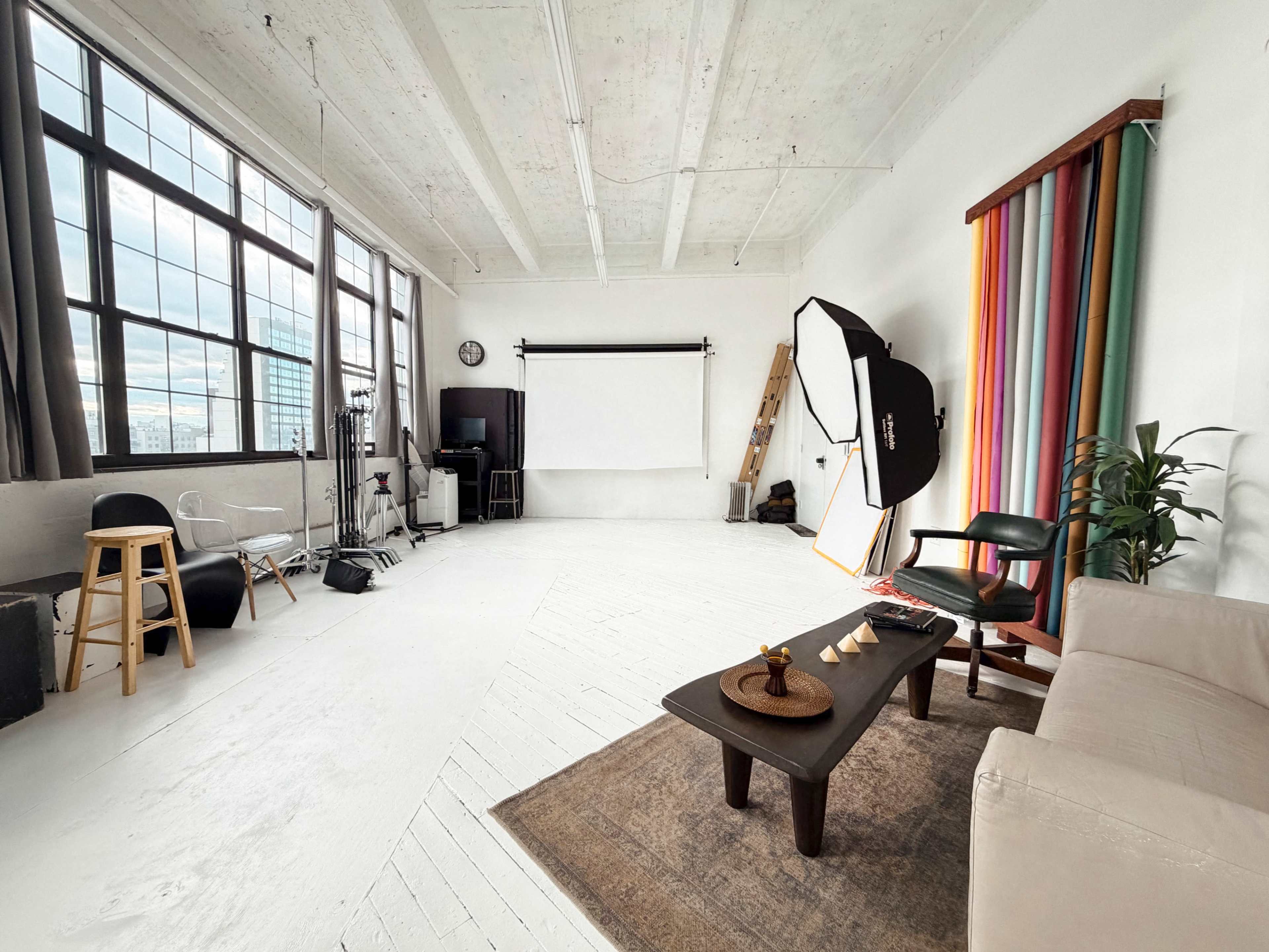 The image shows a bright, spacious photography studio with large windows, various lighting equipment, a seating area, and colorful backdrops.