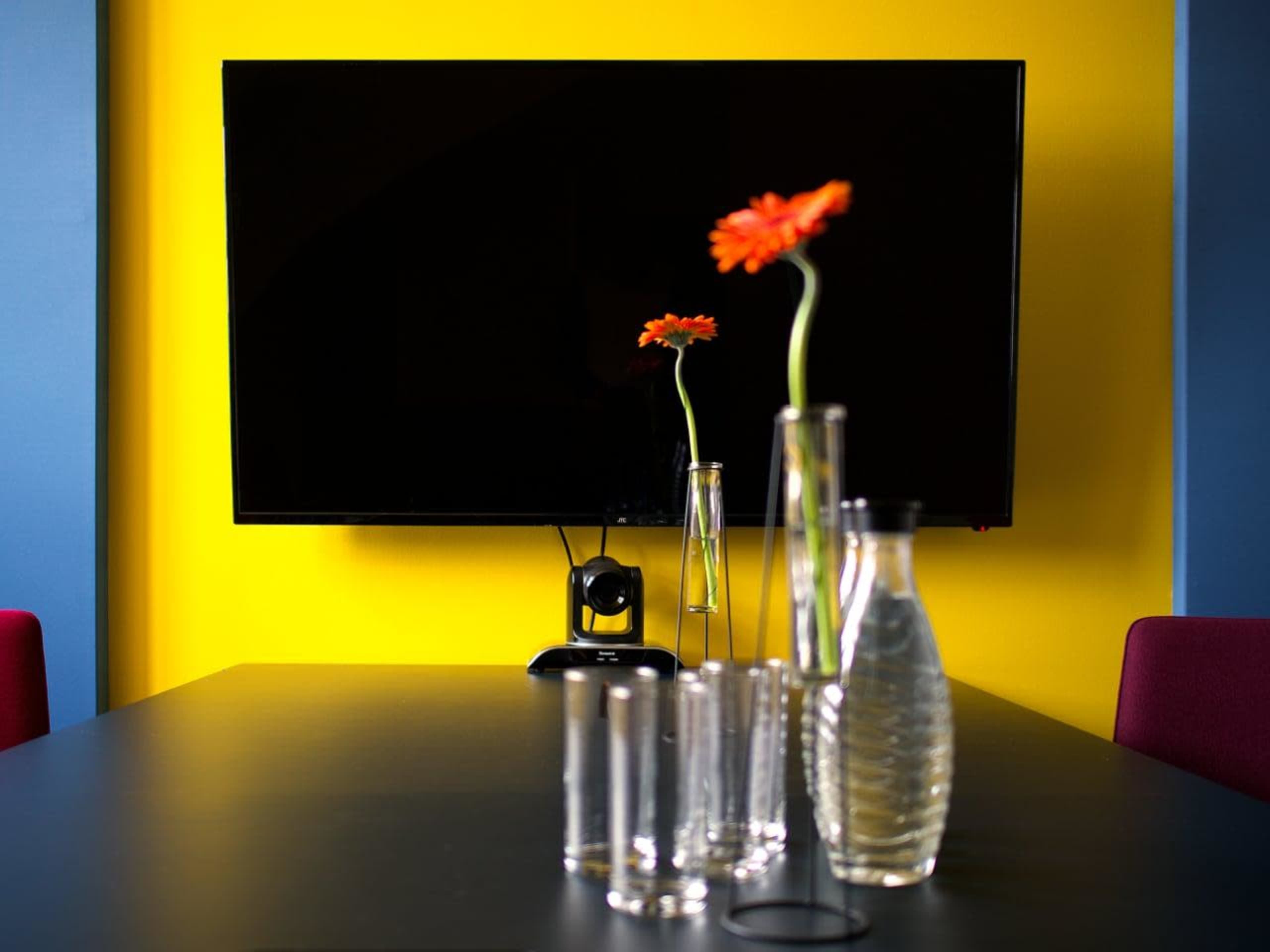 A modern conference room features a large television screen against a bright yellow wall, with several clear glass vases holding flowers arranged on a black table.
