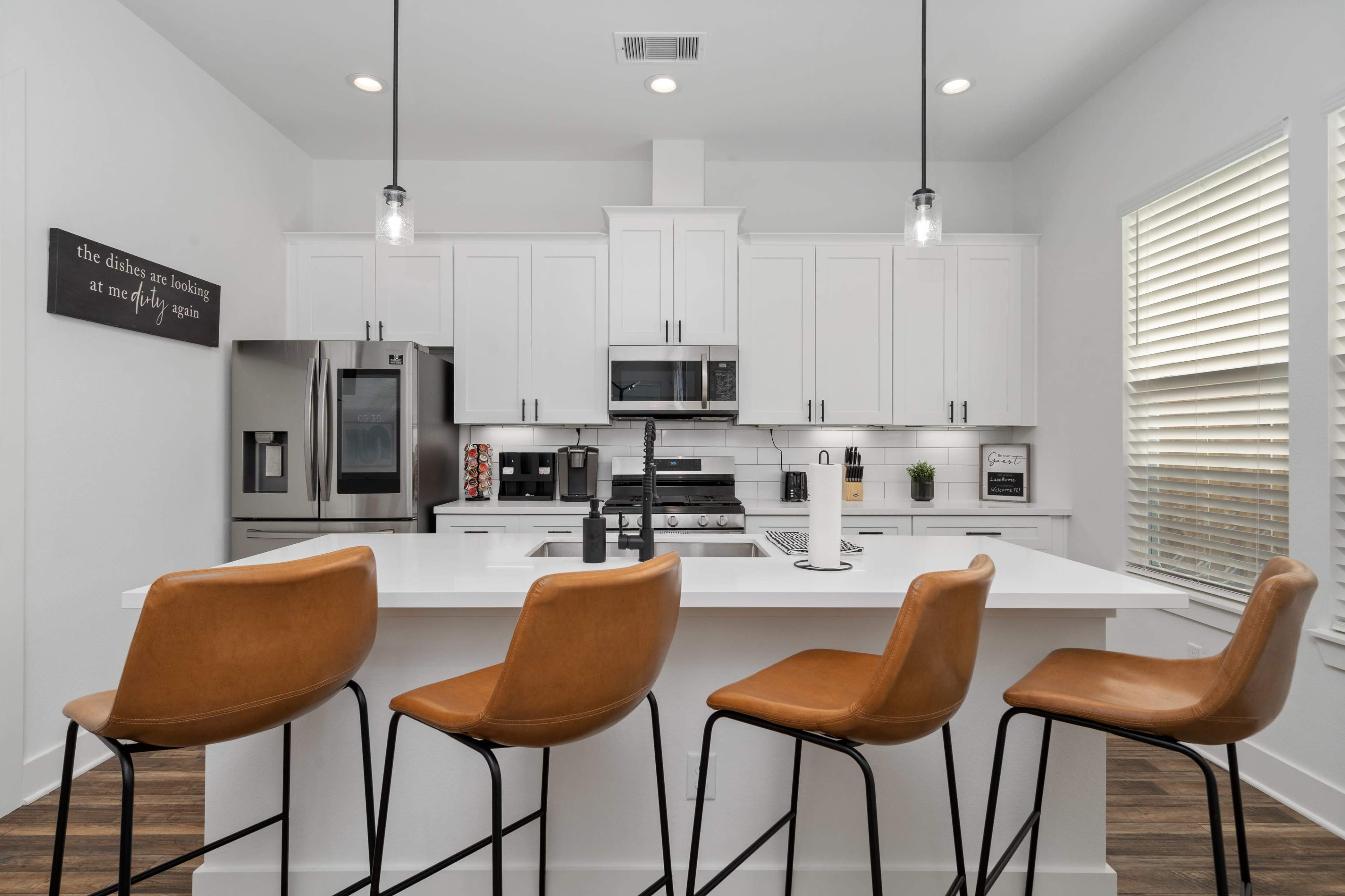 A modern kitchen featuring a white island with four tan bar stools, stainless steel appliances, and bright cabinetry.