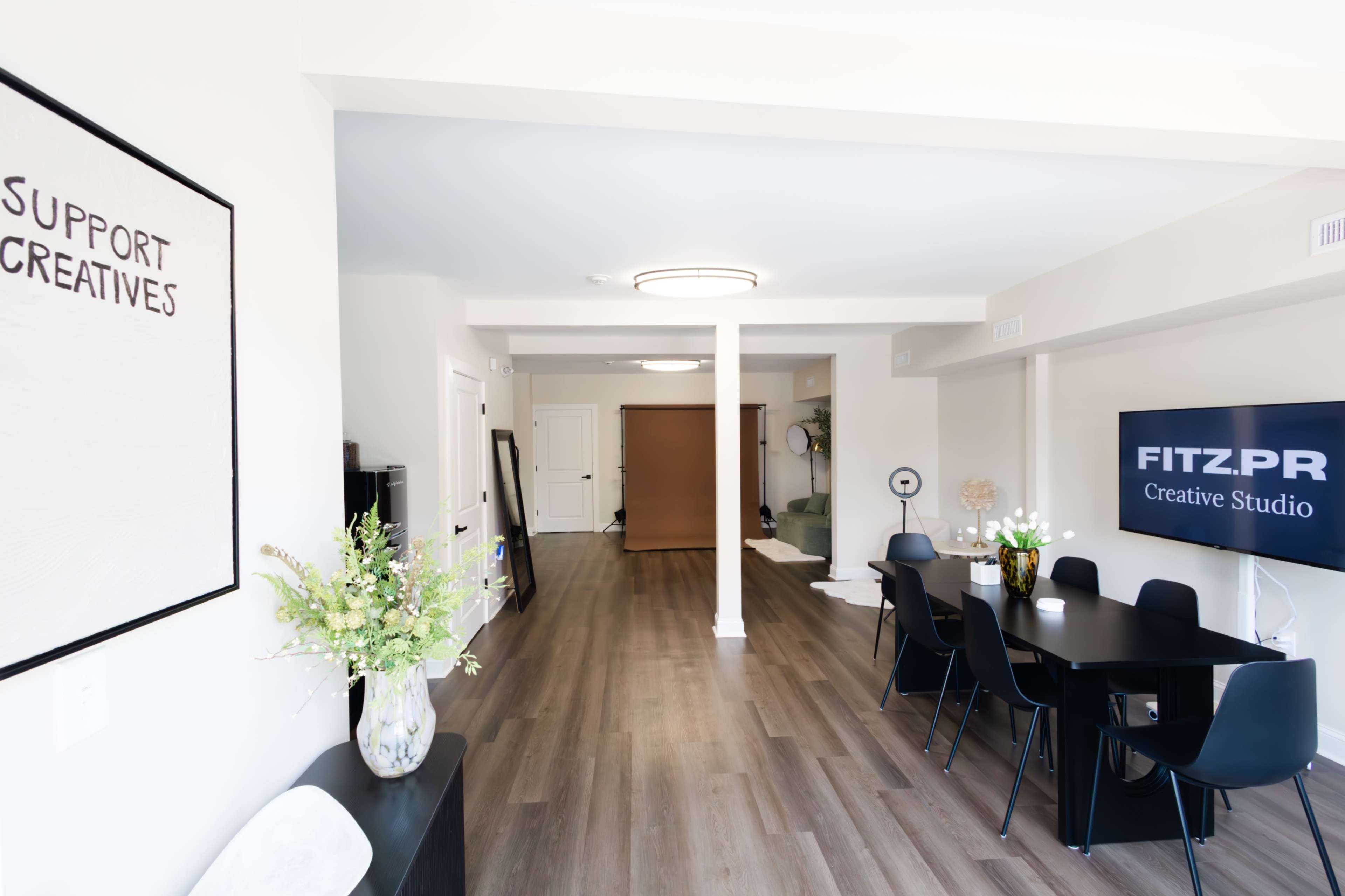 The image shows a modern, open-concept studio space with a dining table and chairs, decorative plants, and a backdrop for creative work.