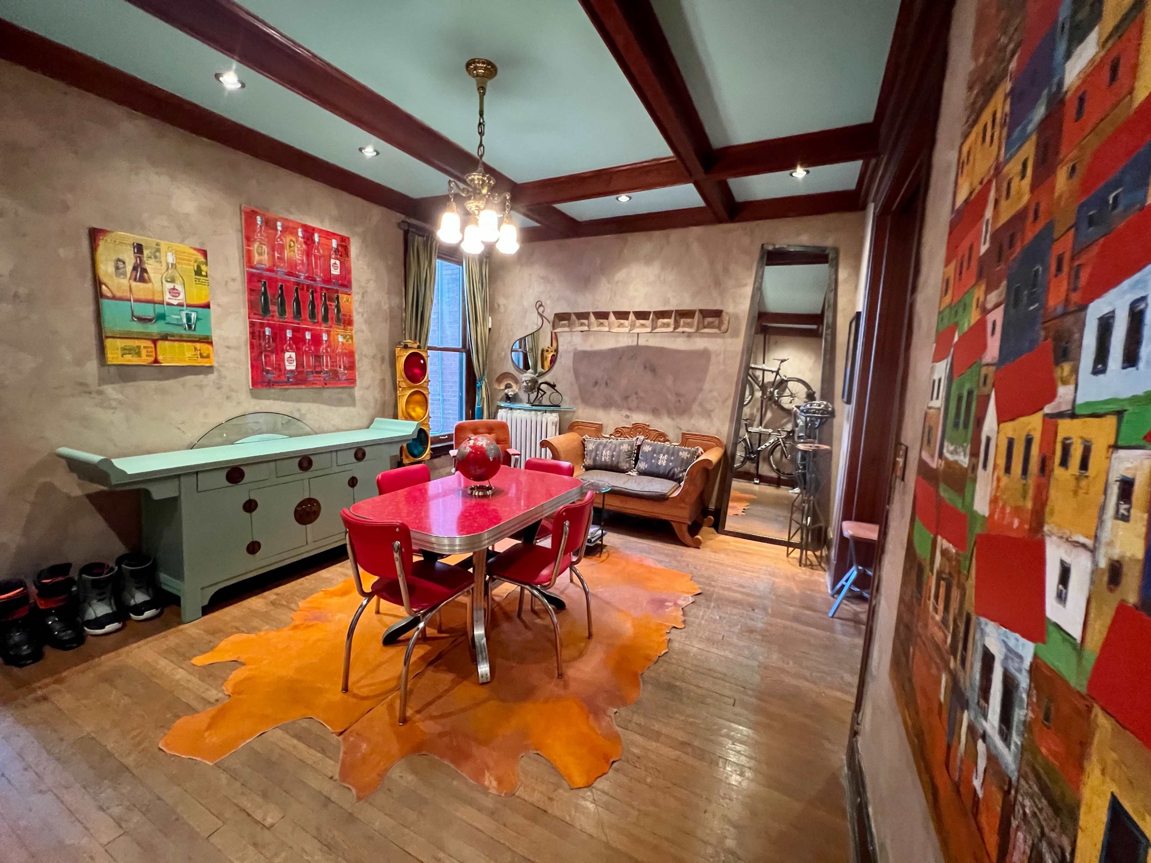 The room features a reddish-orange rug, a dining table surrounded by red chairs, and walls adorned with colorful artwork and various decorations.