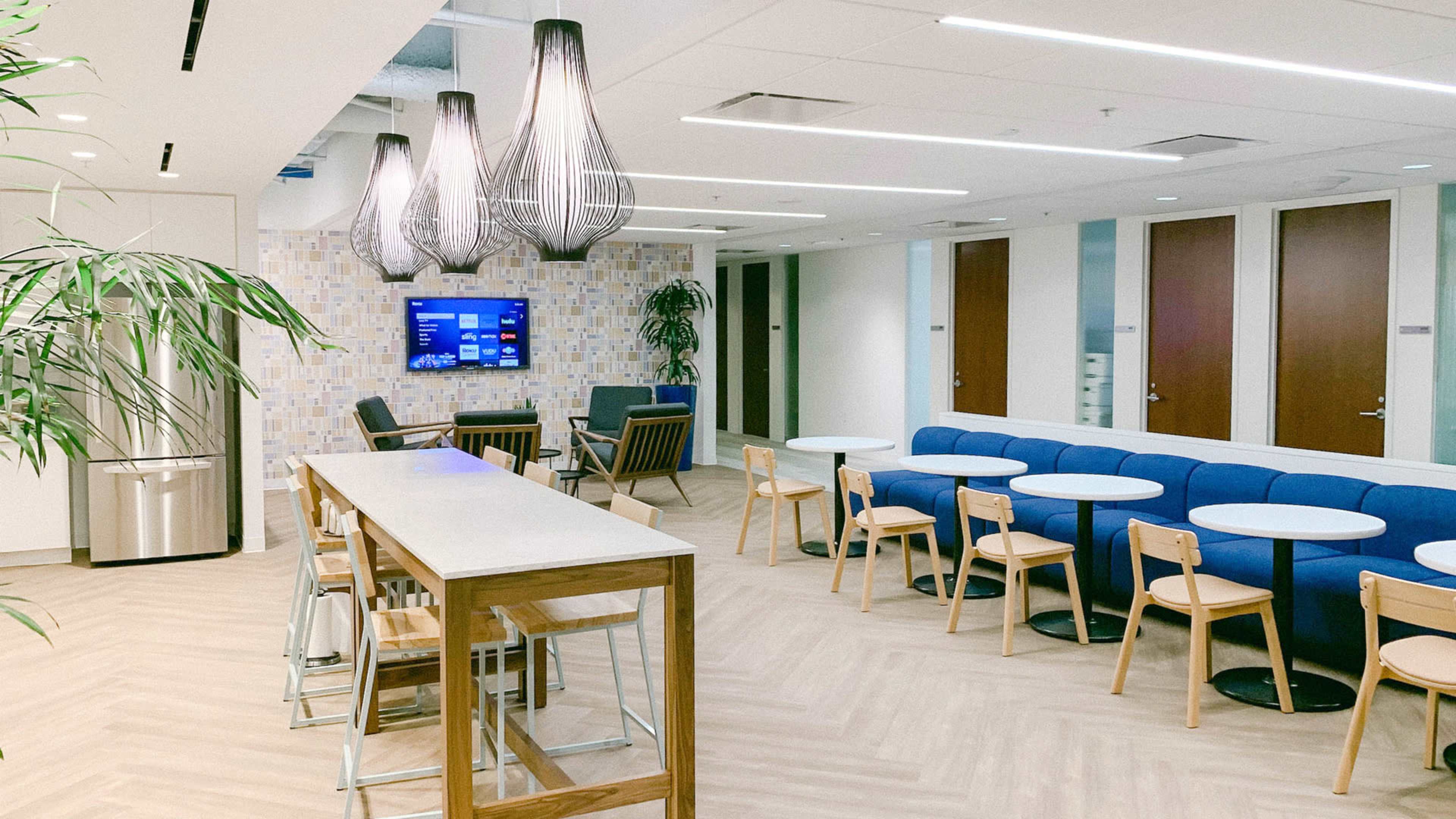 The image shows a modern office break room featuring a long wooden bar, several round tables with chairs, a blue sofa, a television on the wall, and plants in the corners.