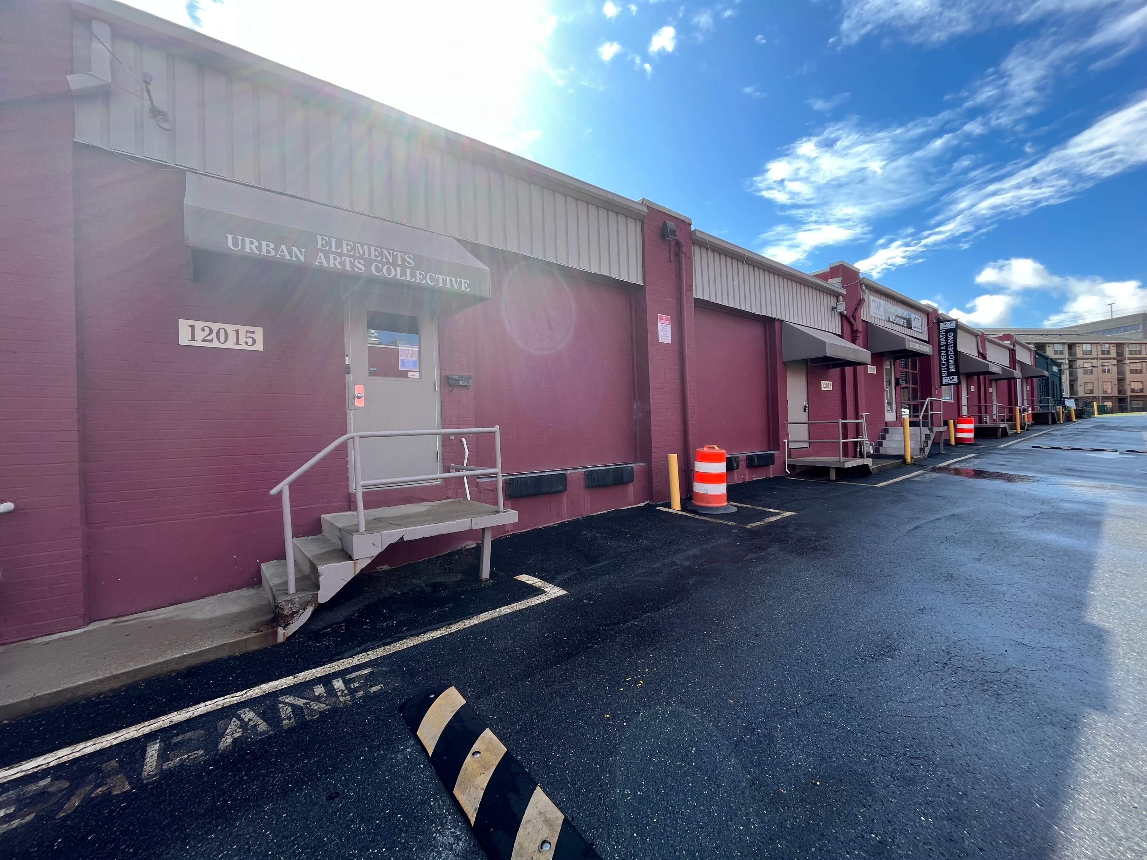 A red brick building labeled "Elements Urban Arts Collective" with loading docks and a clear blue sky in the background.