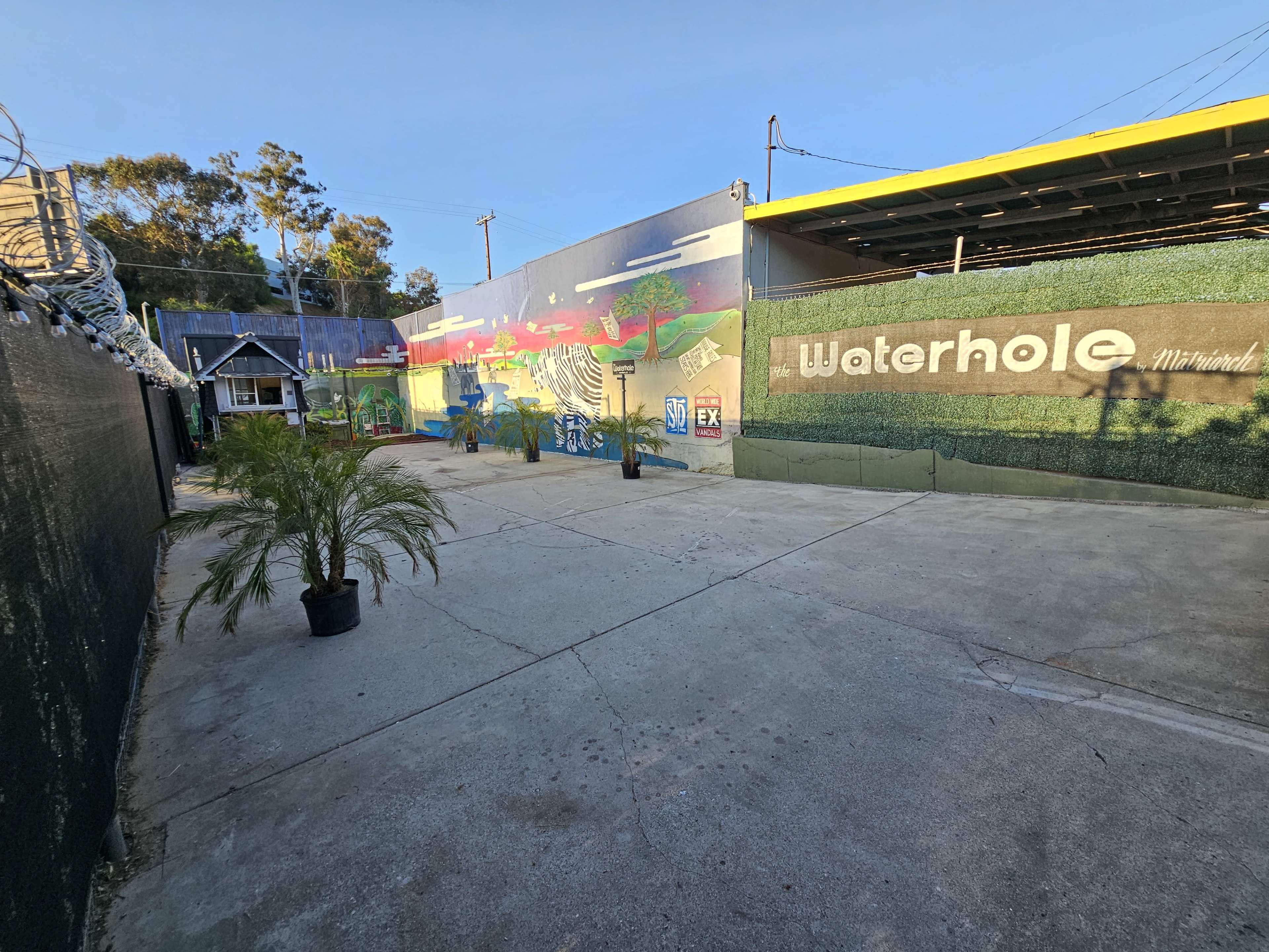 A spacious outdoor area featuring a mural on a wall labeled "Waterhole," with concrete flooring and potted plants.