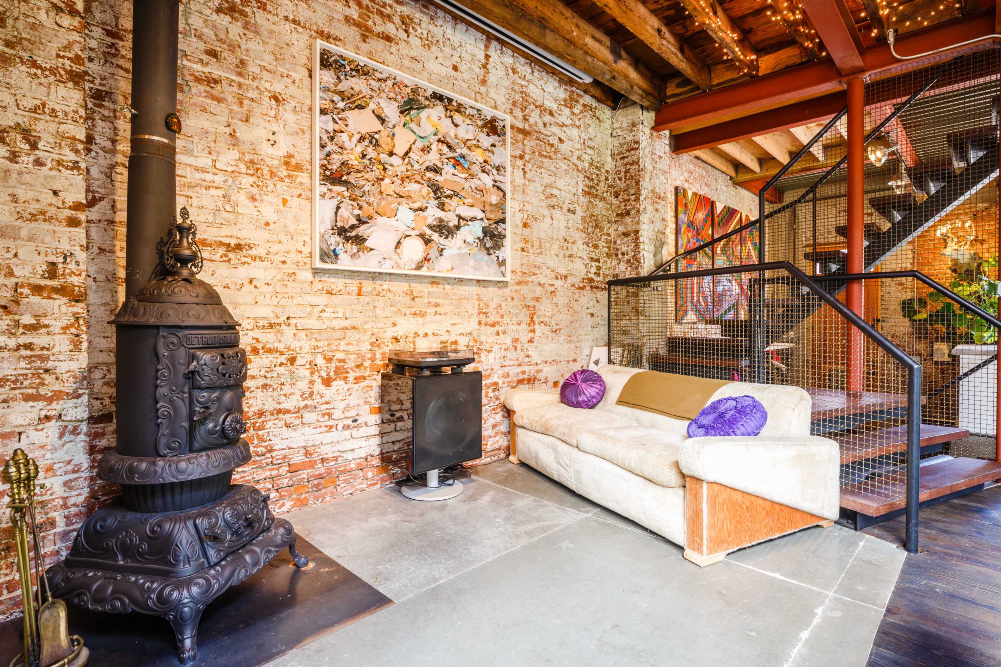 The image shows a cozy living space with a vintage cast iron stove, a cream-colored sofa adorned with decorative pillows, and exposed brick walls, along with art pieces in a loft-like setting.