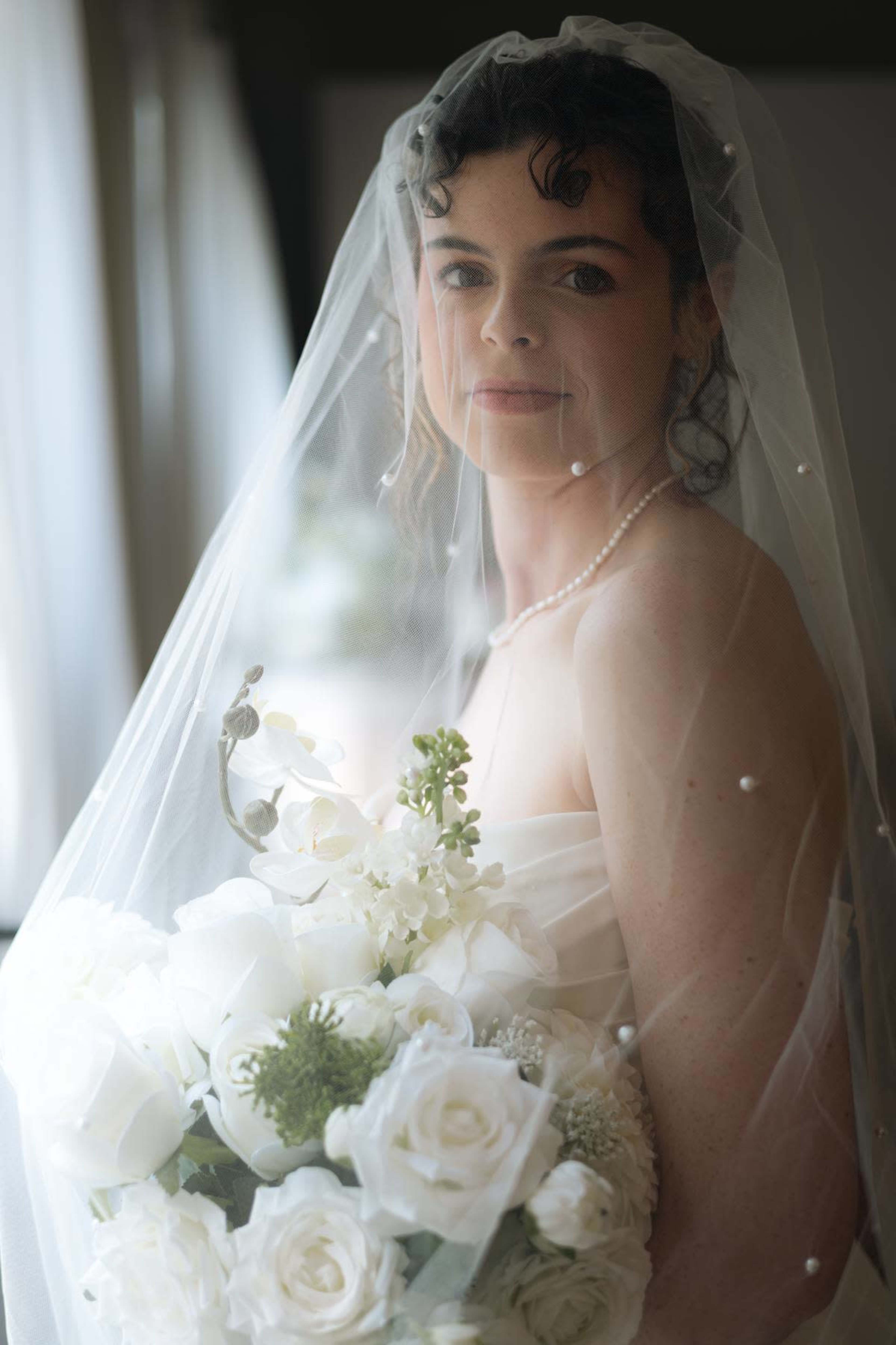 A bride in a white dress poses with a bouquet of white flowers while wearing a veil and pearl necklace.