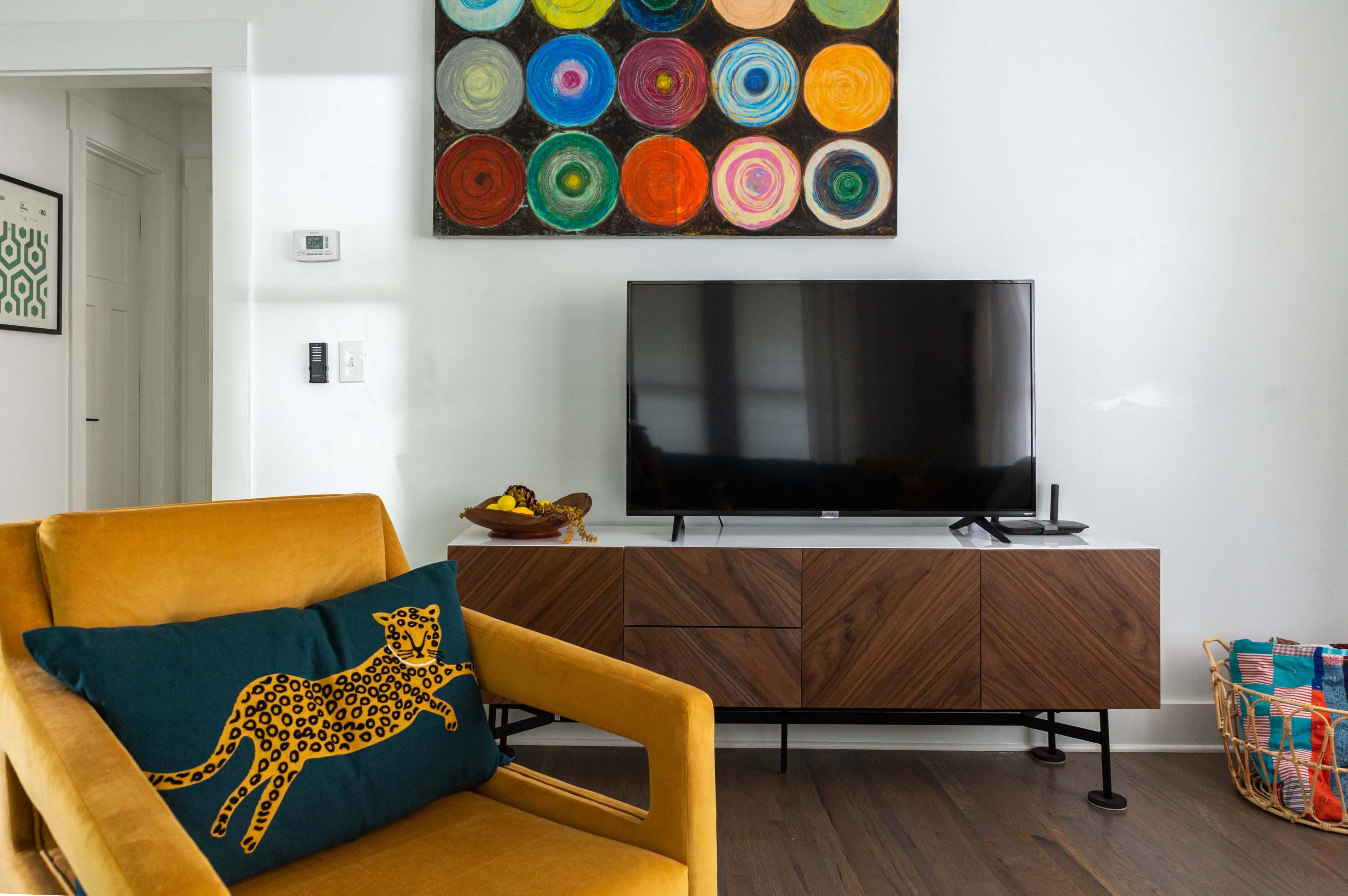 A modern living room features a yellow armchair with a decorative pillow, a wooden TV stand, and a colorful circular painting on the wall above a flat-screen television.