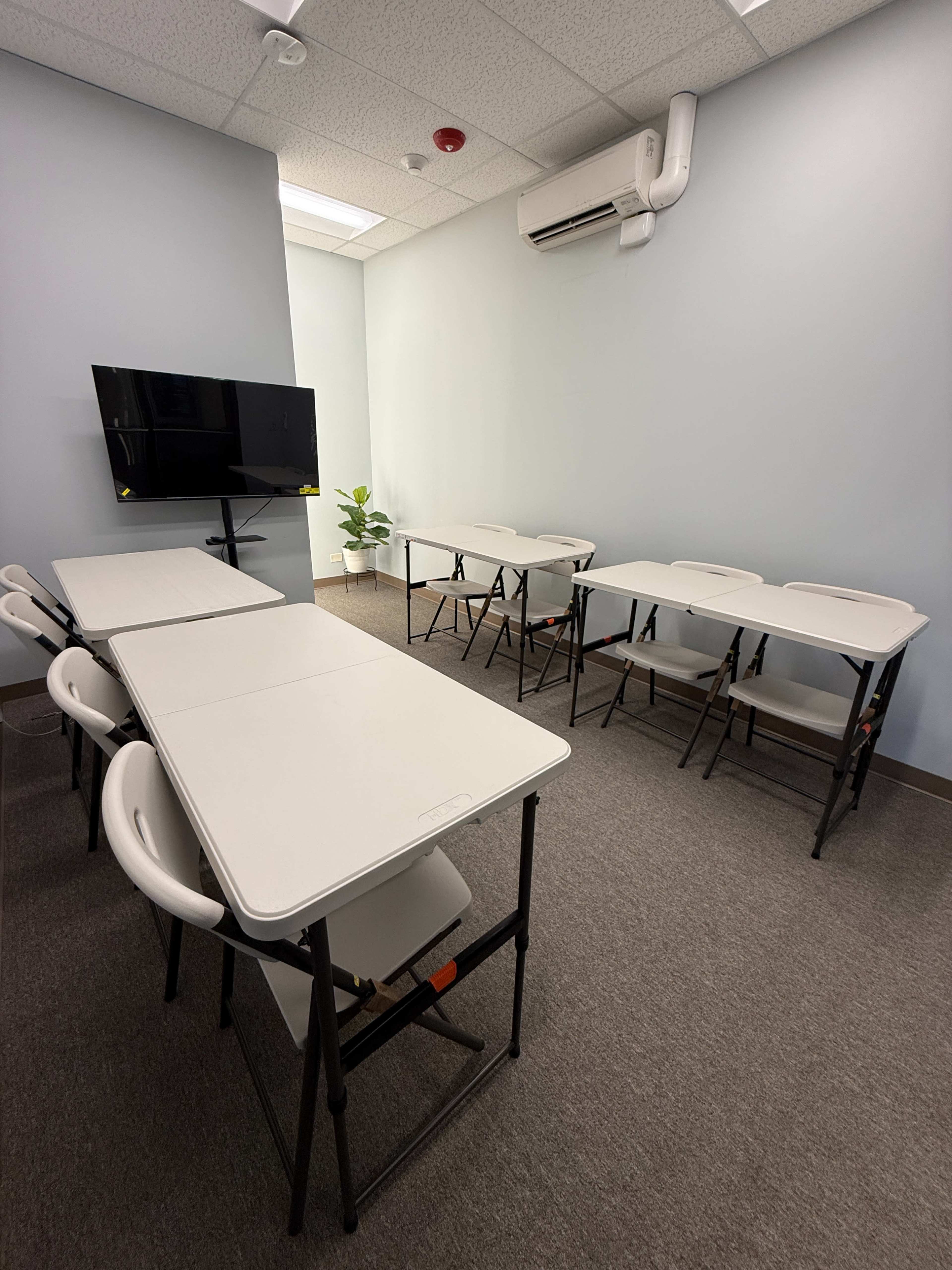 The image shows a small, empty classroom with white folding tables arranged in two rows facing a wall-mounted television and a potted plant in the corner.