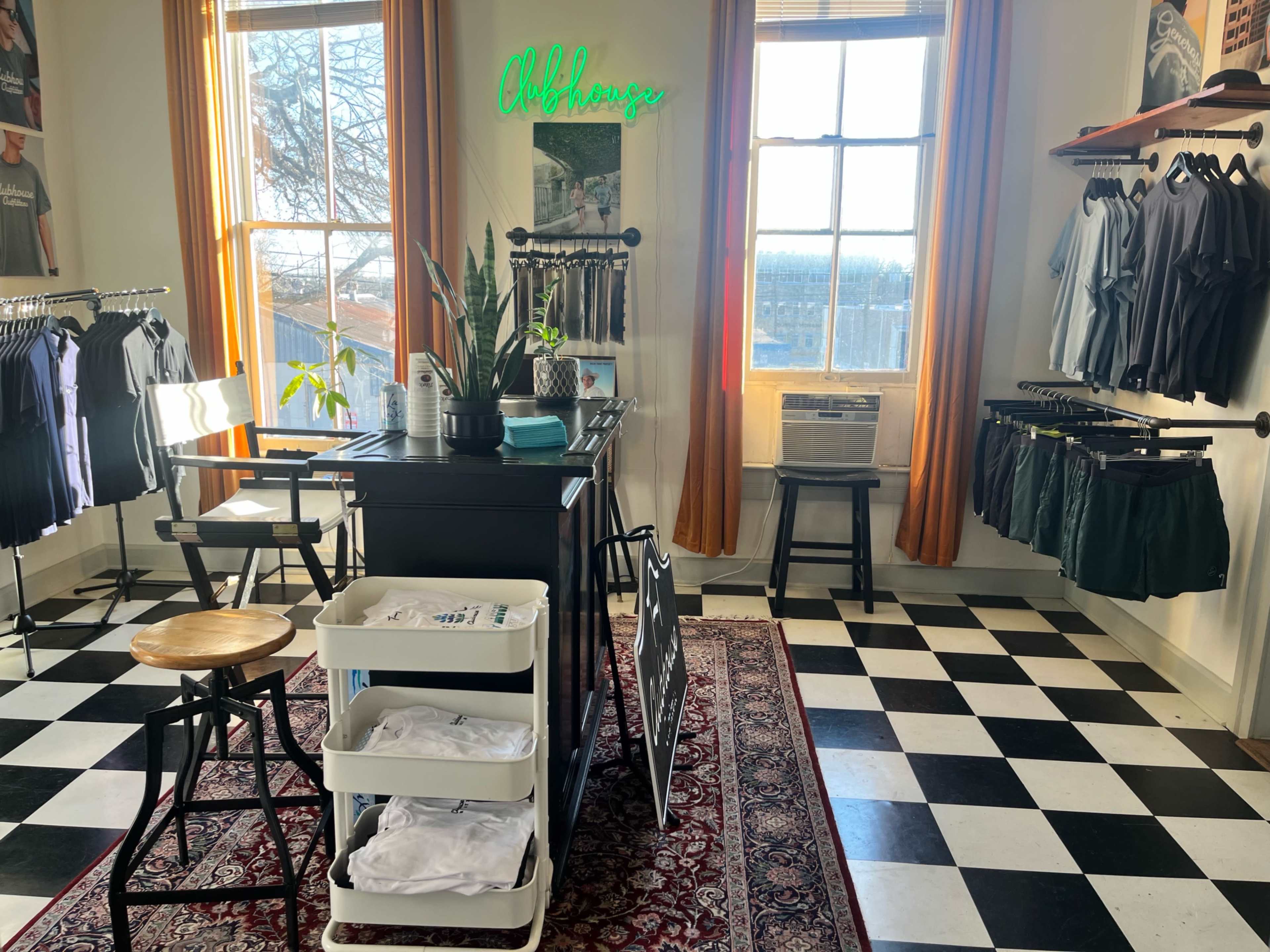 The image shows a retail store featuring a black-and-white checkered floor, clothing racks displaying various garments, and a counter with a neon sign in a well-lit room.
