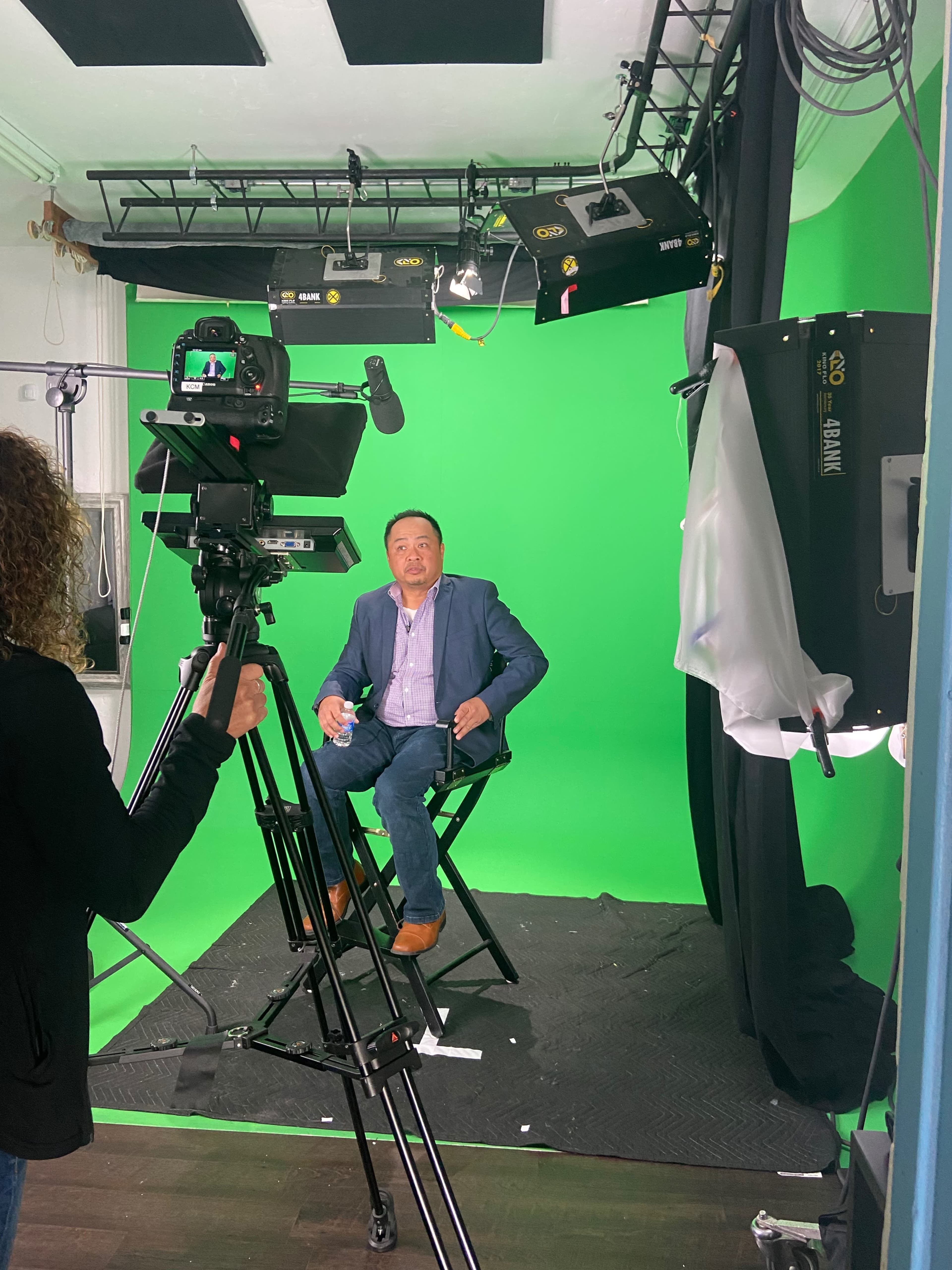 A man in a blue suit sits in a director's chair in front of a green screen while being filmed by a camera operator.