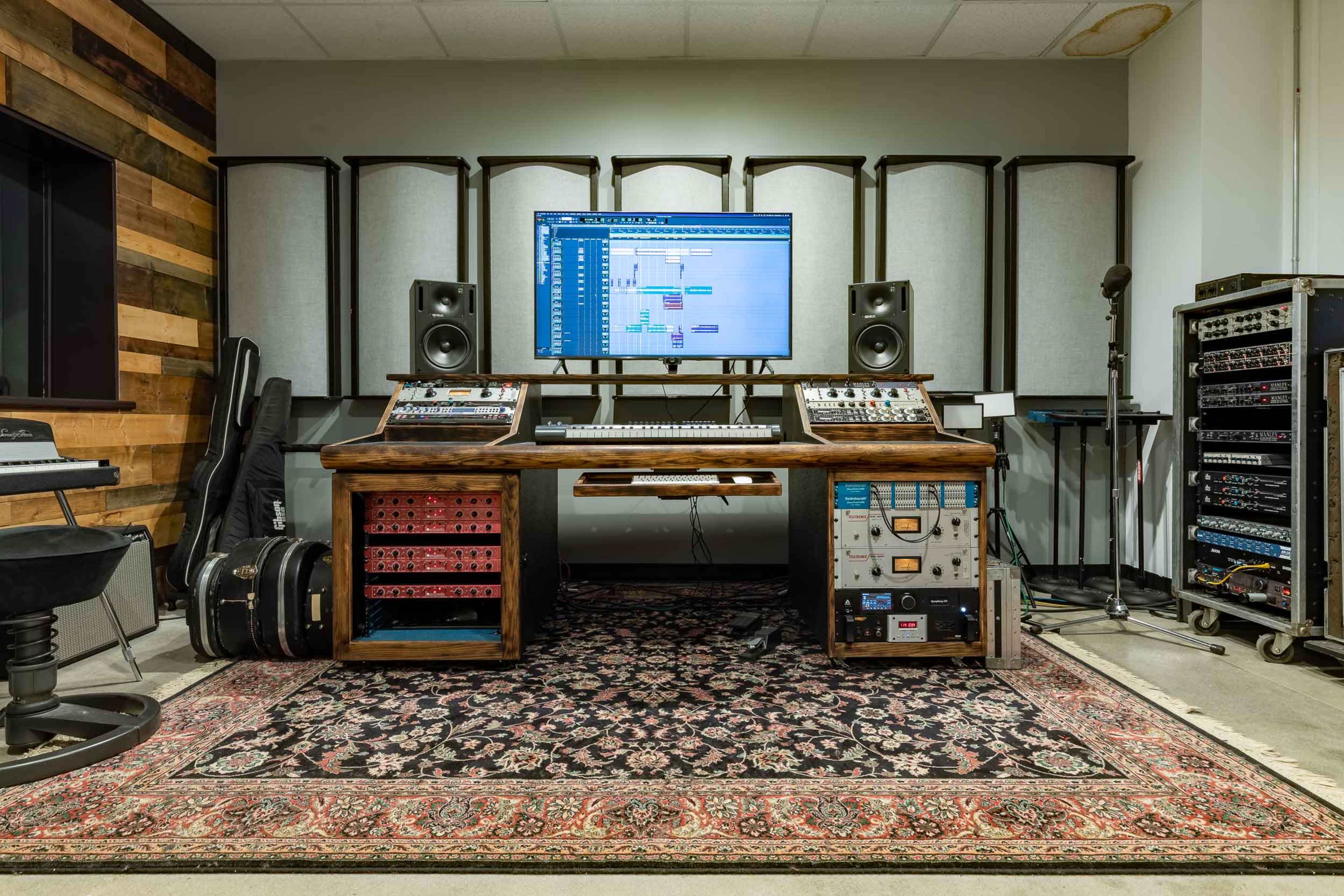 A music production studio features a large wooden desk with audio equipment, a computer display, and surrounding soundproof panels.