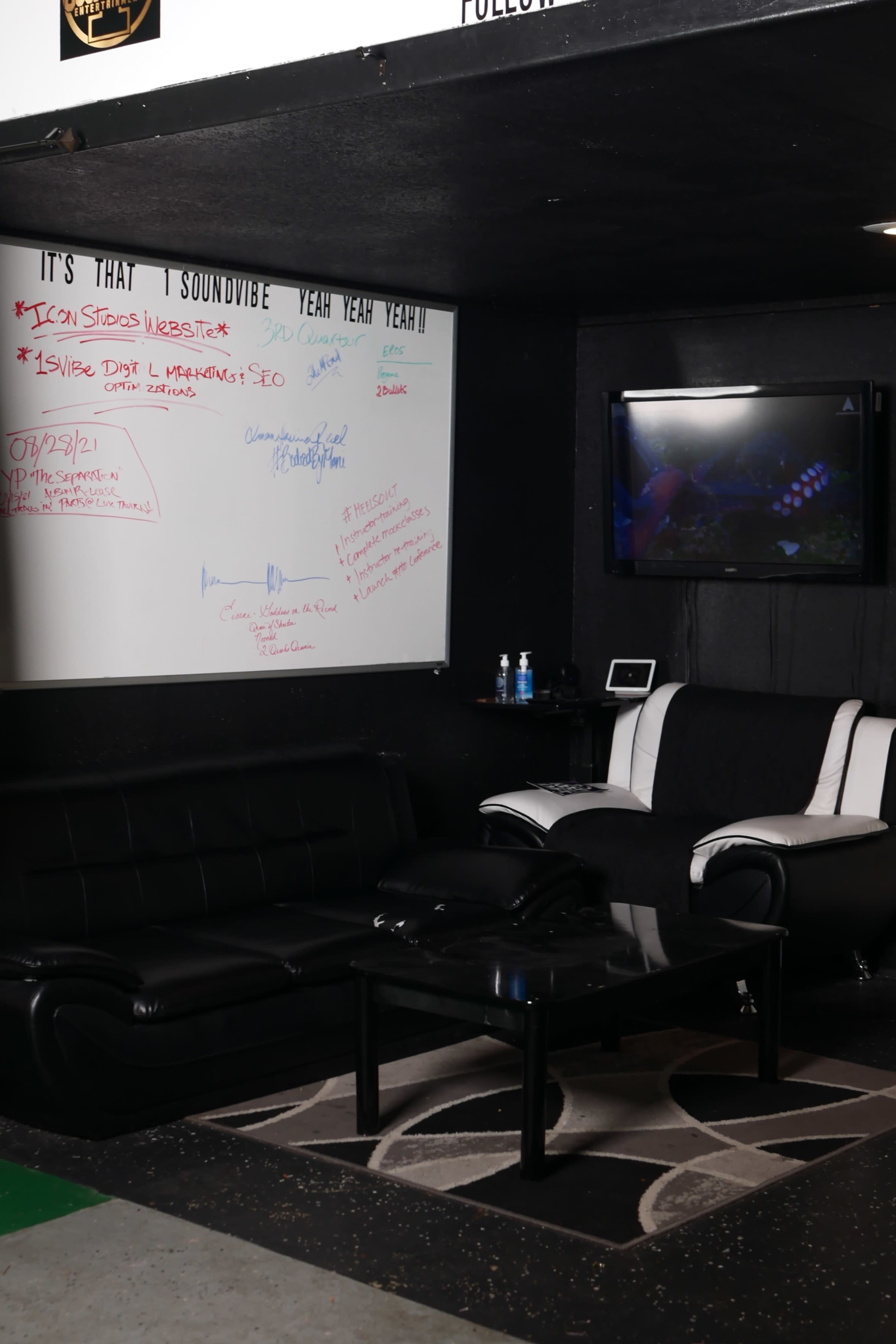 The image shows a small lounge area with black furniture, a glass coffee table, a whiteboard covered in colorful notes and sketches, and a television mounted on the wall.