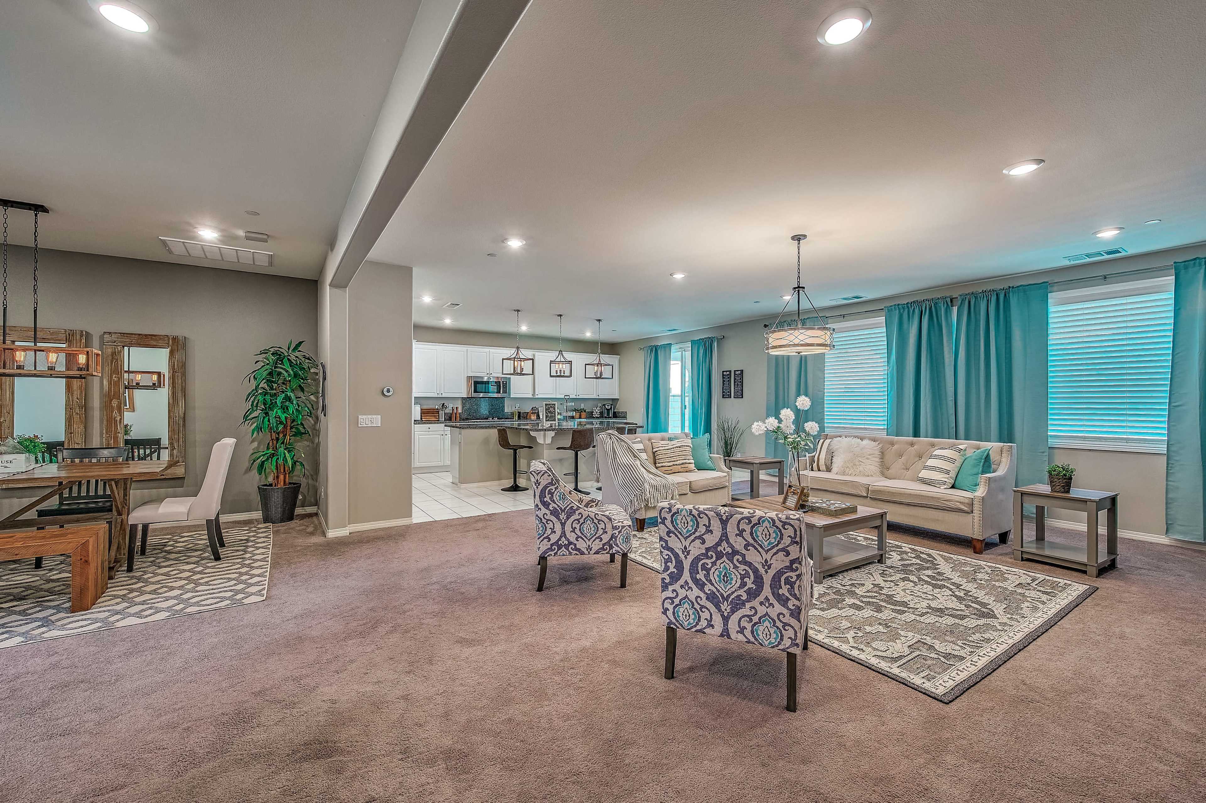 The image shows an open-concept living area featuring a cozy seating arrangement, a dining table, and a kitchen in the background, all illuminated by natural light.