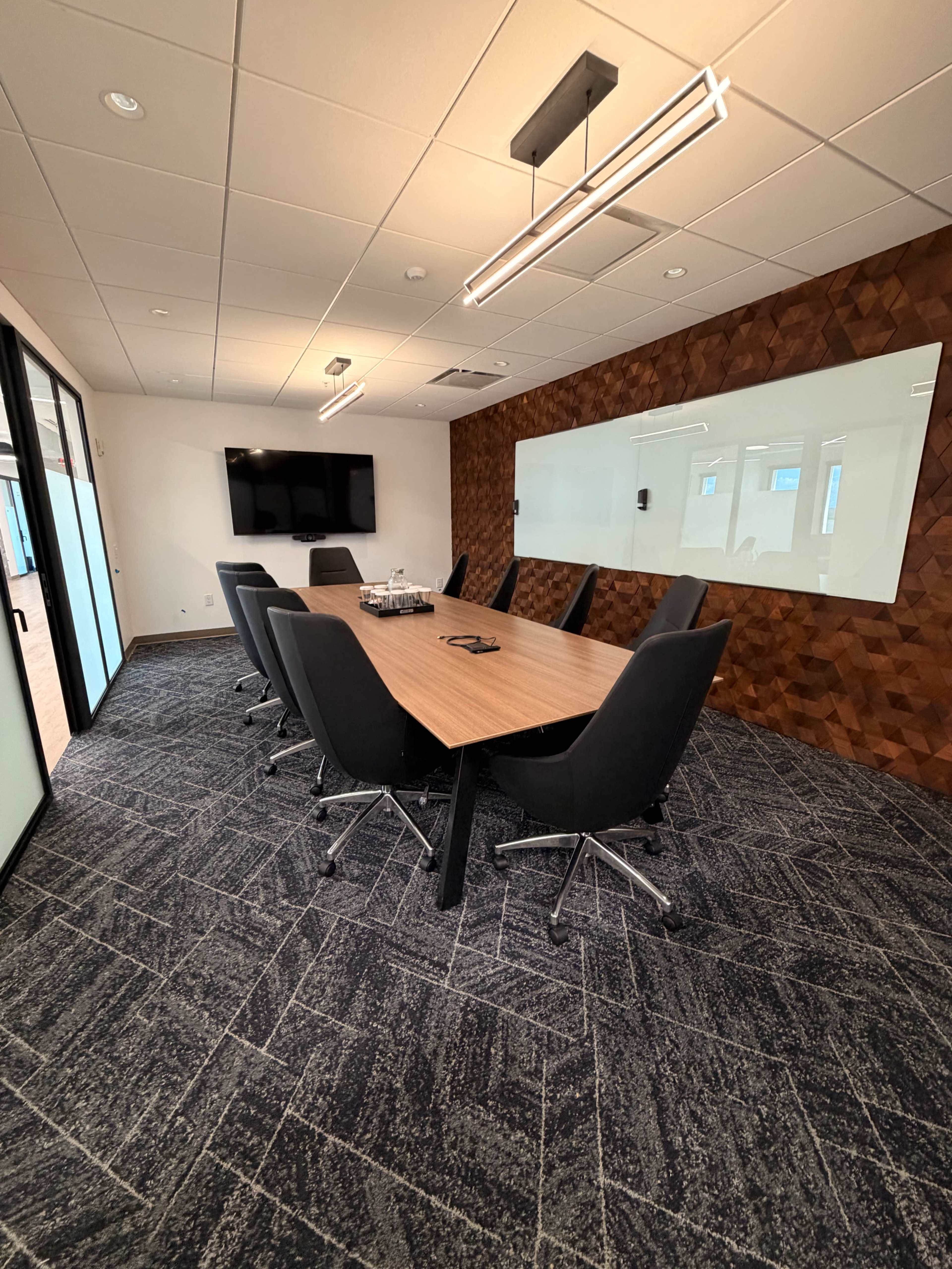 The image shows a modern conference room featuring a large wooden table surrounded by black chairs, with a flat-screen TV mounted on the wall and a glass whiteboard on another wall.