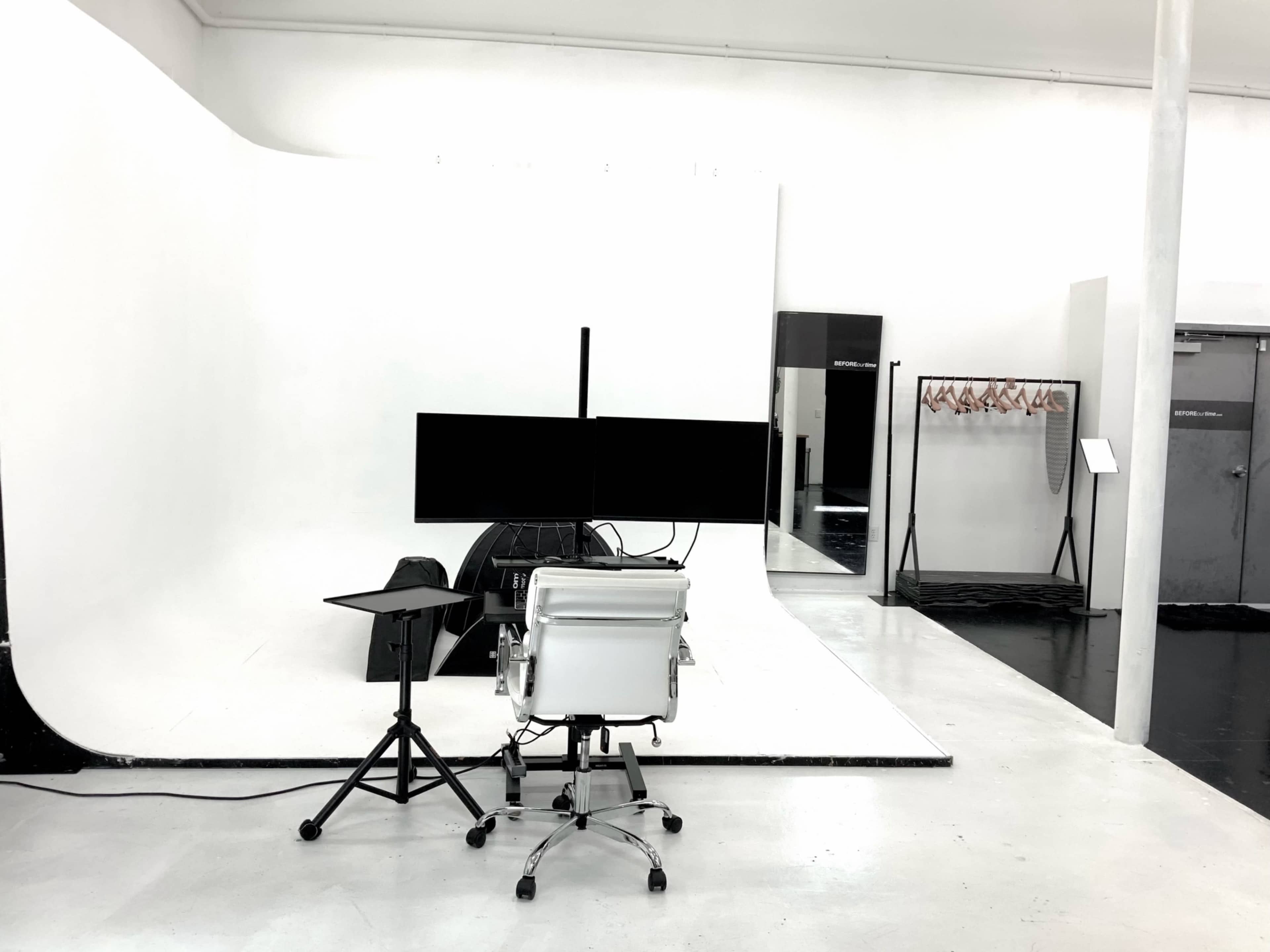 The image shows a minimalist studio setup with a white backdrop, a chair with multiple monitors, and a small table on a polished floor.