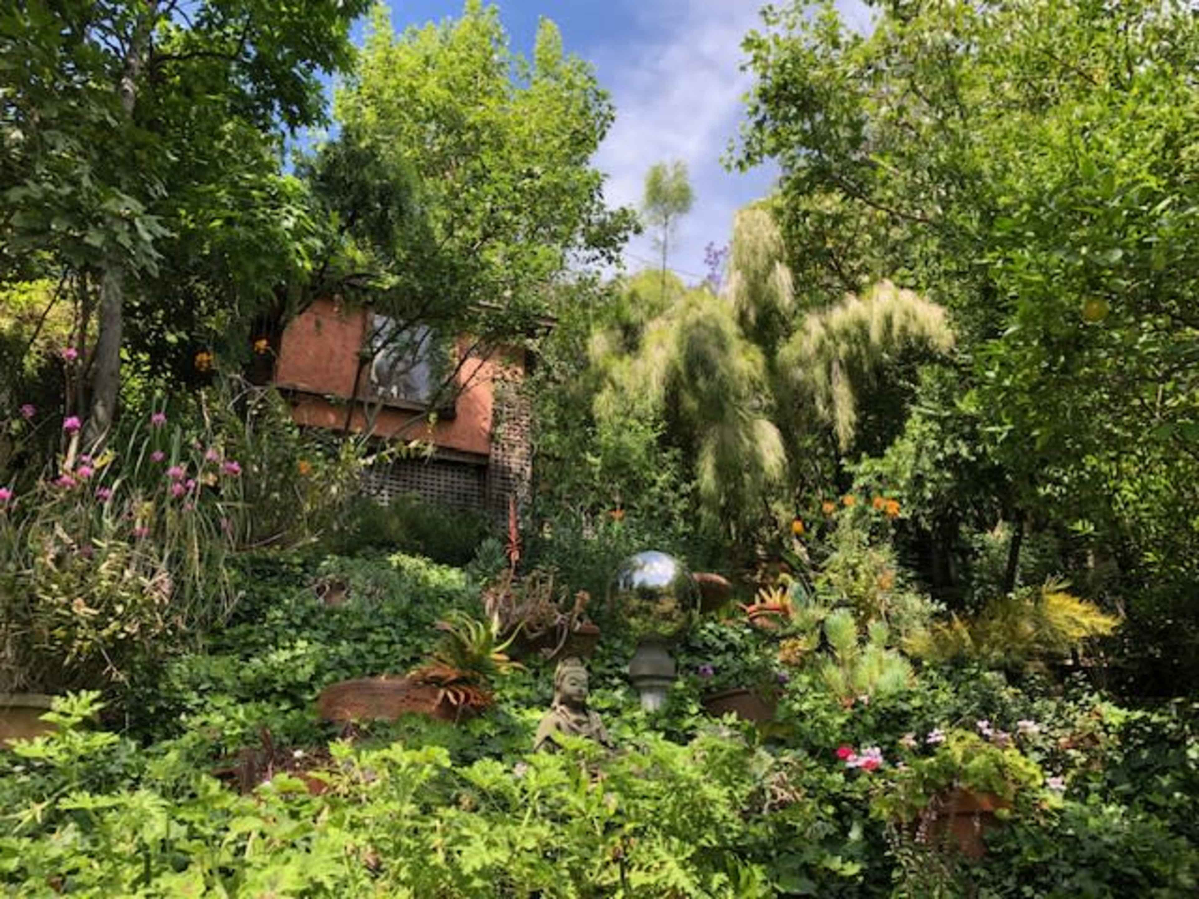 A lush garden features a variety of plants, sculptures, and a reflective orb set against a backdrop of trees and a building.