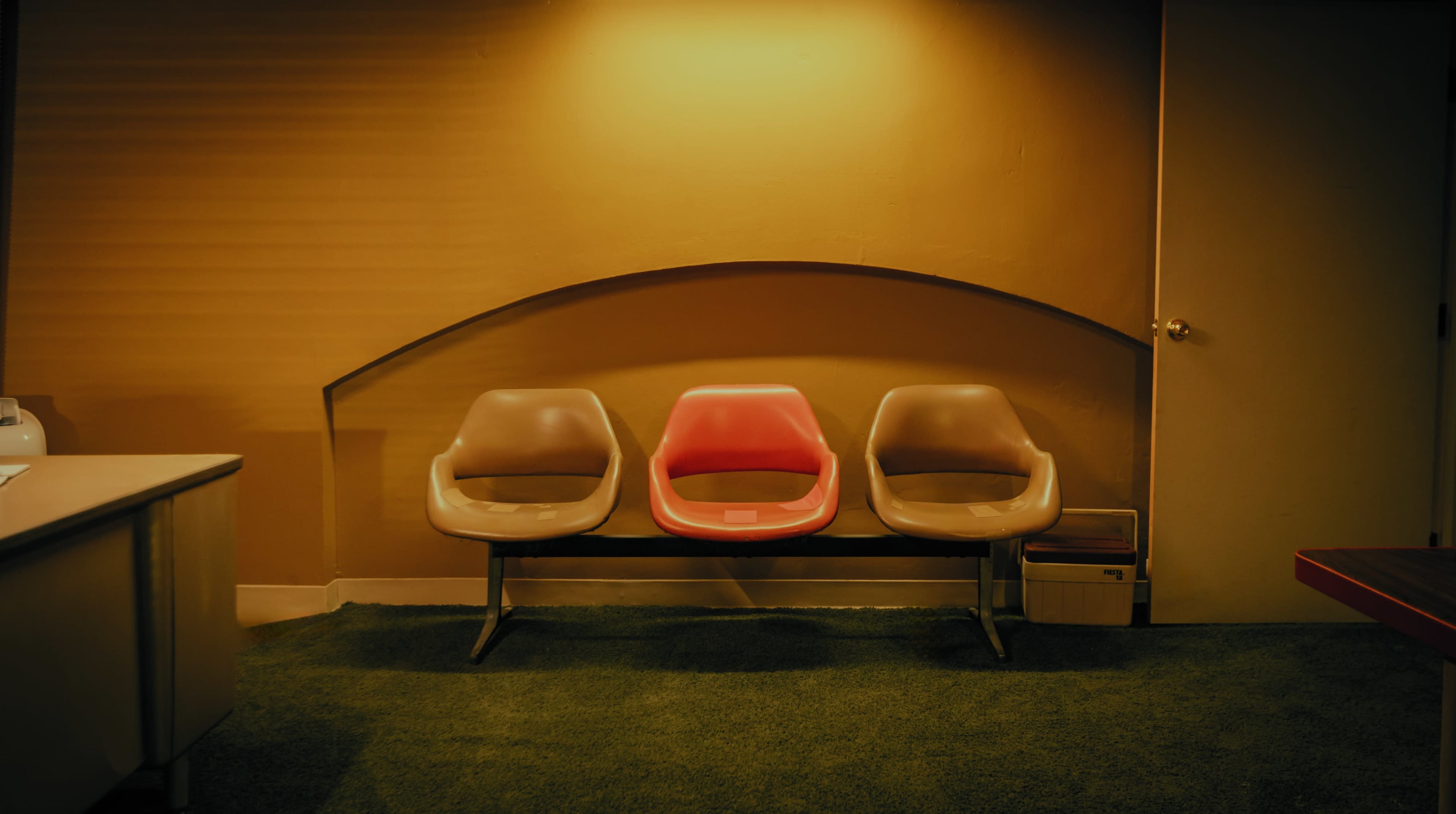 A row of three chairs, with the center chair being red, is positioned against a wall in a dimly lit room.