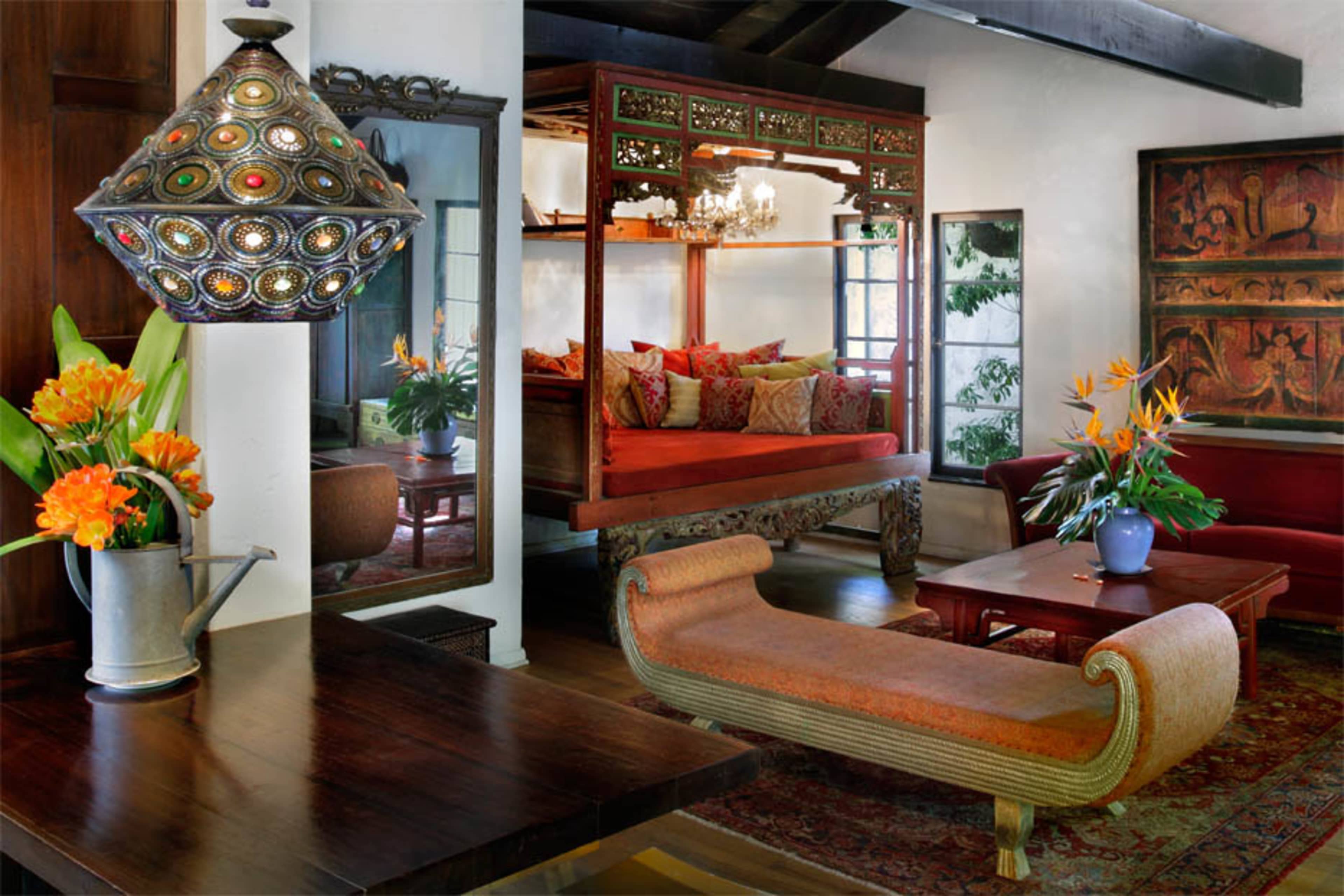 A brightly decorated living room features a large, ornate bed, a wooden table with floral arrangements, and various pieces of art on the walls.