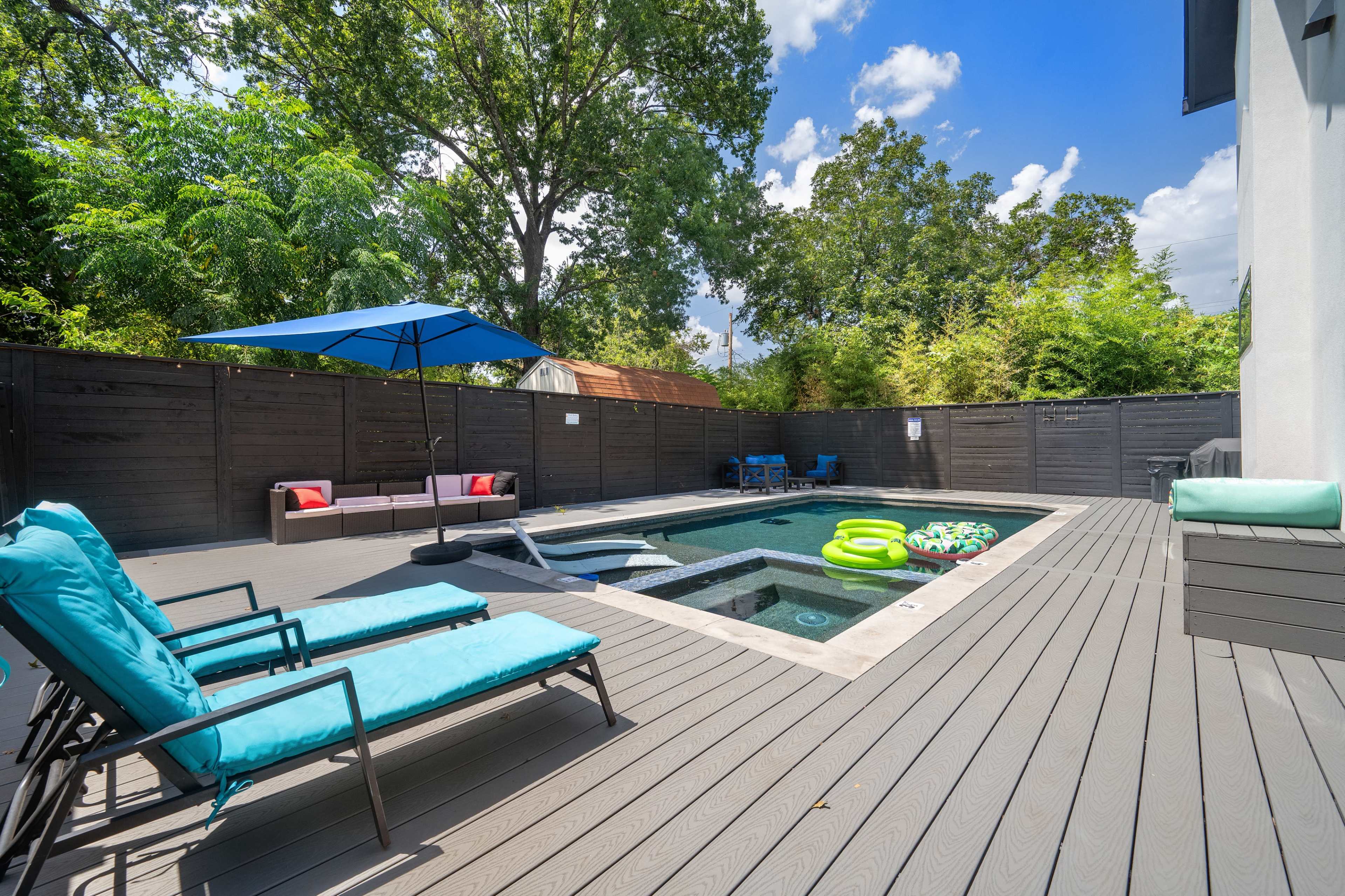 The image shows a modern outdoor space featuring a swimming pool surrounded by lounge chairs, a blue umbrella, and various floatation devices on a deck.