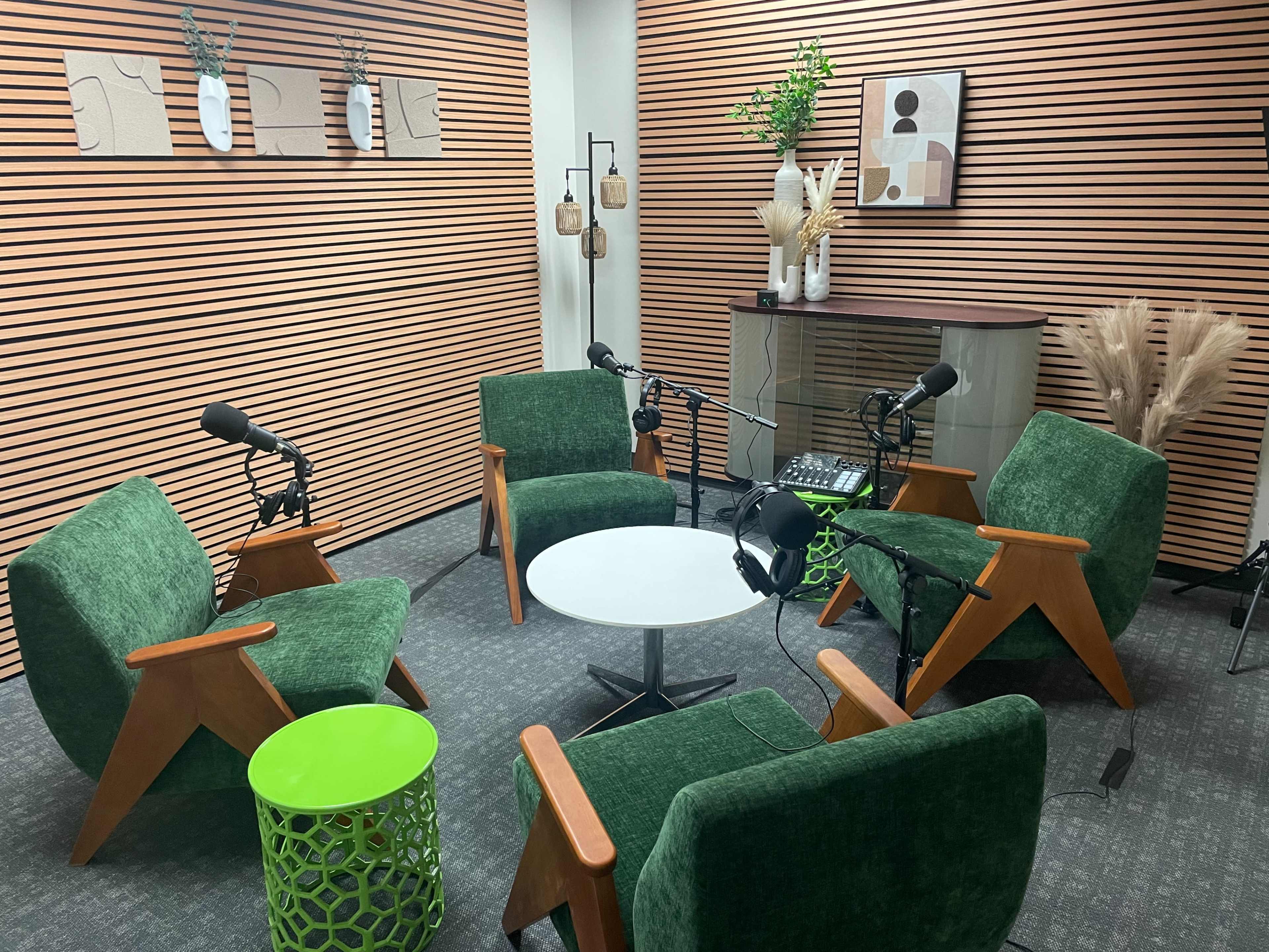 The image shows a podcast recording studio featuring four green chairs, a round white table, and audio equipment, with wooden paneling and decorative plants on the walls.