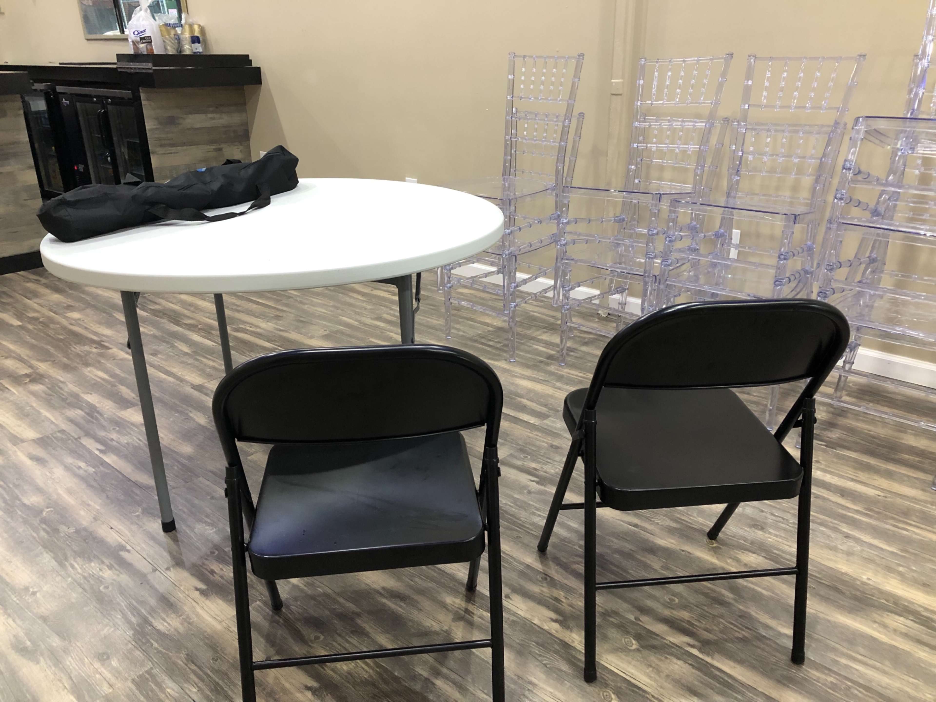 A round white table is positioned in front of two black folding chairs, with clear plastic chairs stacked in the background.
