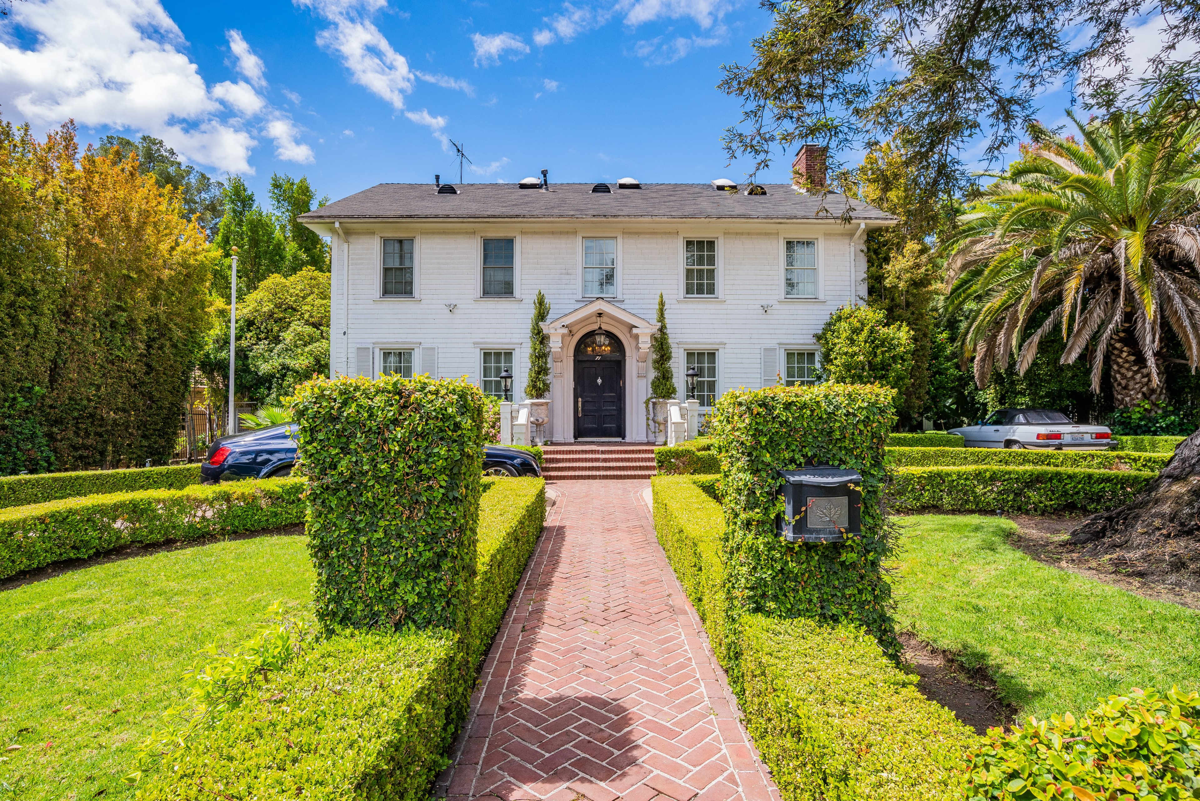 A two-story white house with a brick pathway leading to the front door, flanked by manicured hedges and palm trees.