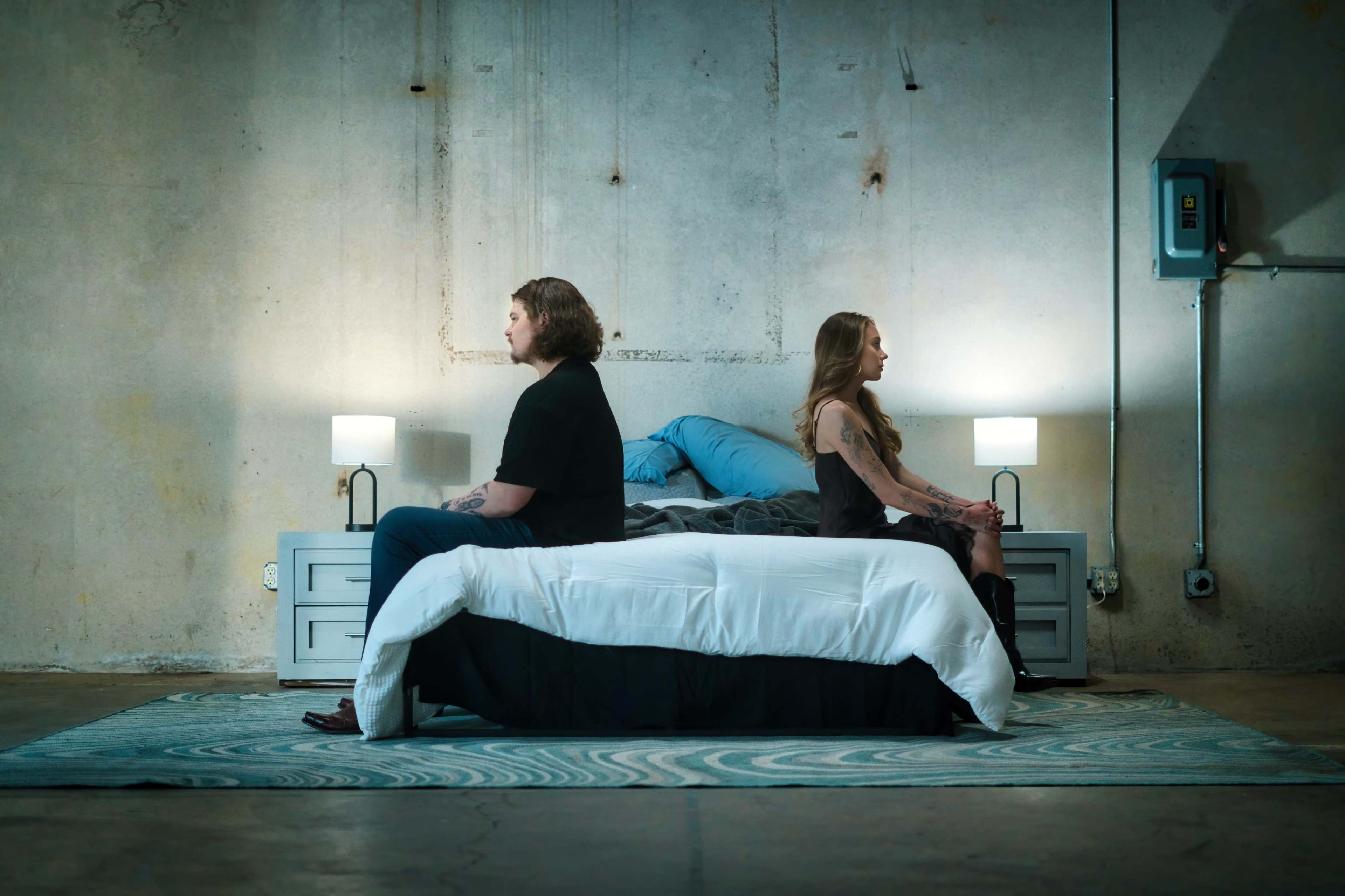A man and a woman sit on opposite sides of a bed in a dimly lit room with concrete walls.