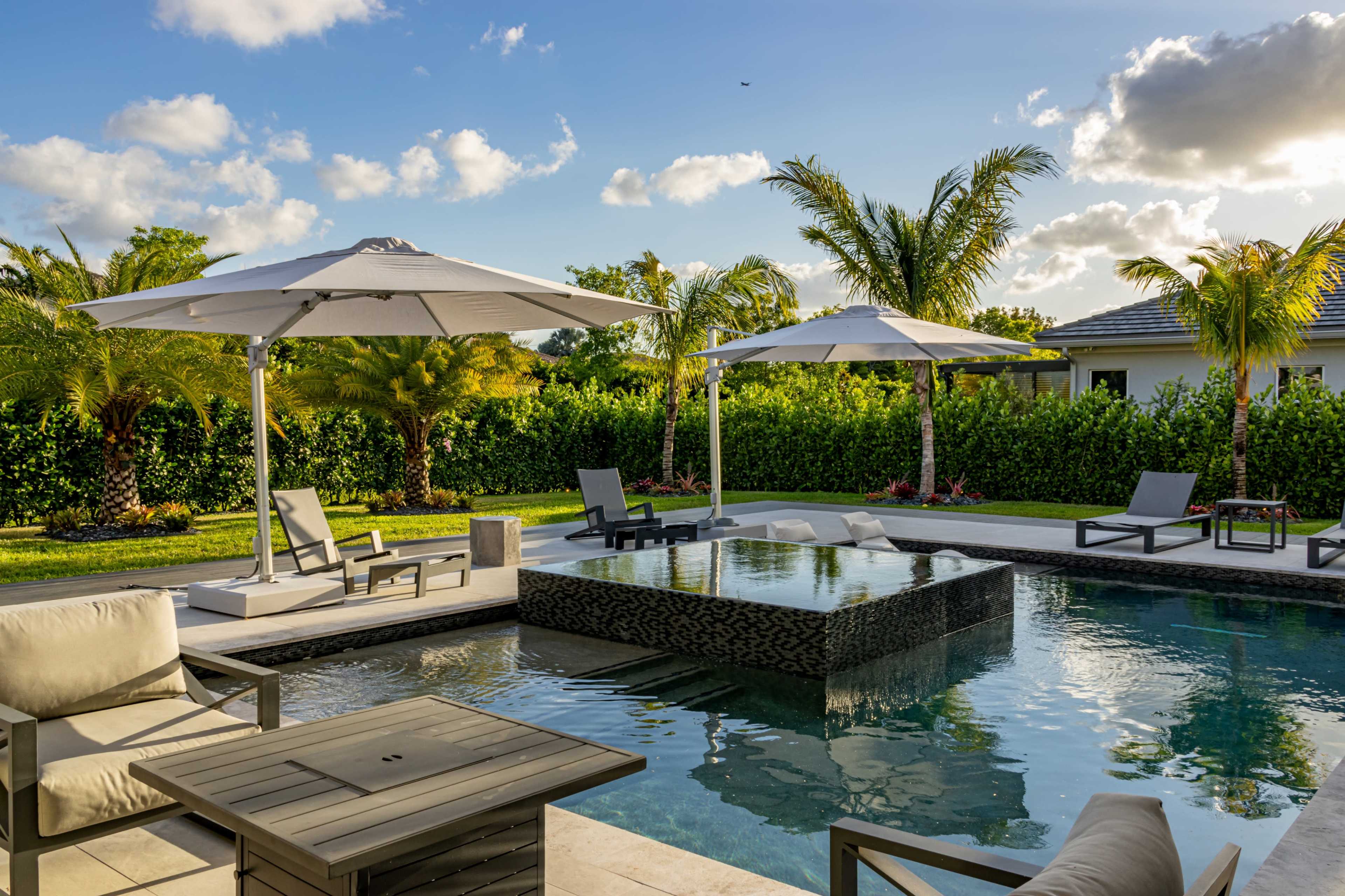The image shows a modern swimming pool area with umbrellas, lounge chairs, and lush greenery in the background.