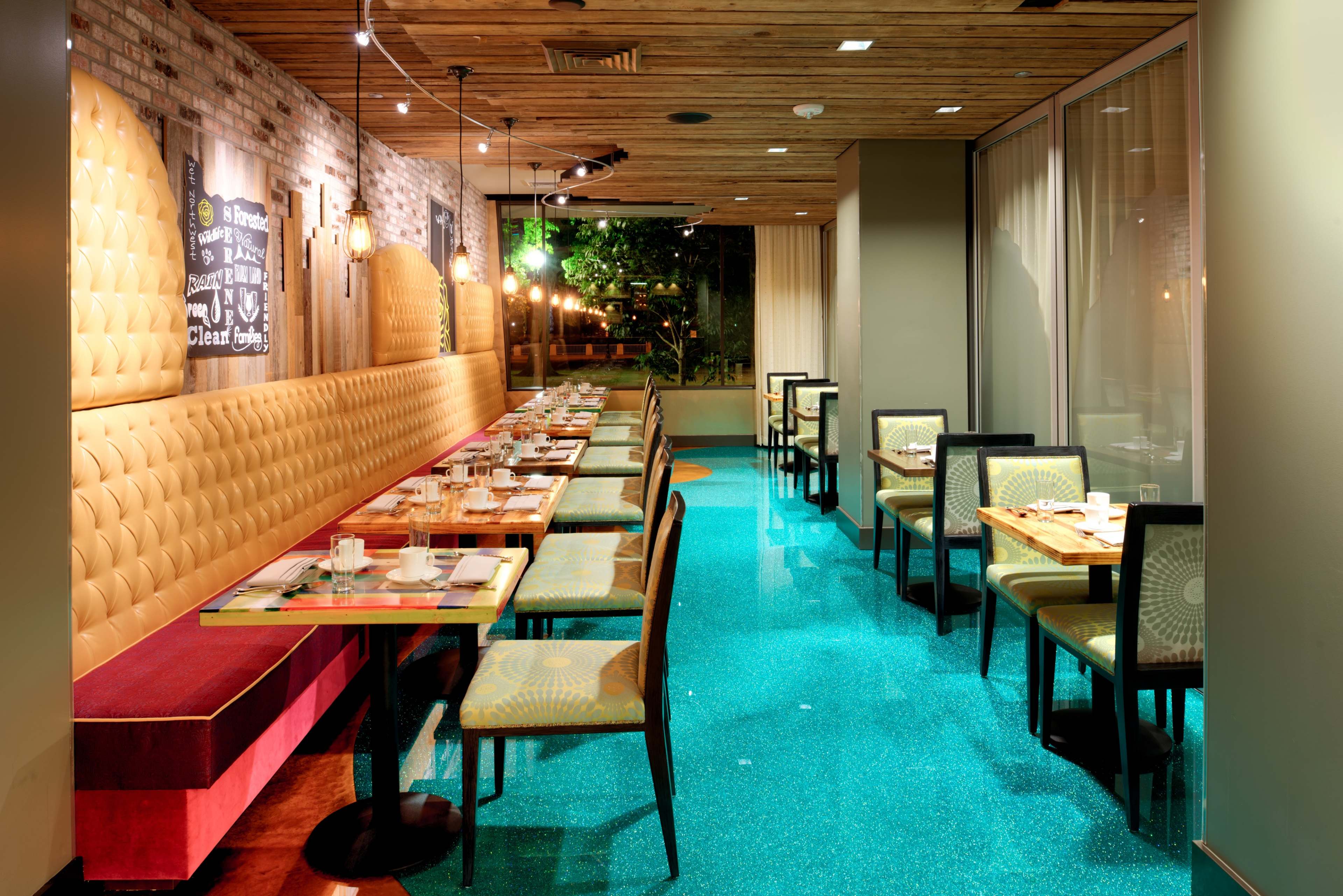 A modern restaurant interior featuring a booth along one side, small tables with chairs arranged throughout, and a turquoise, glittering floor.