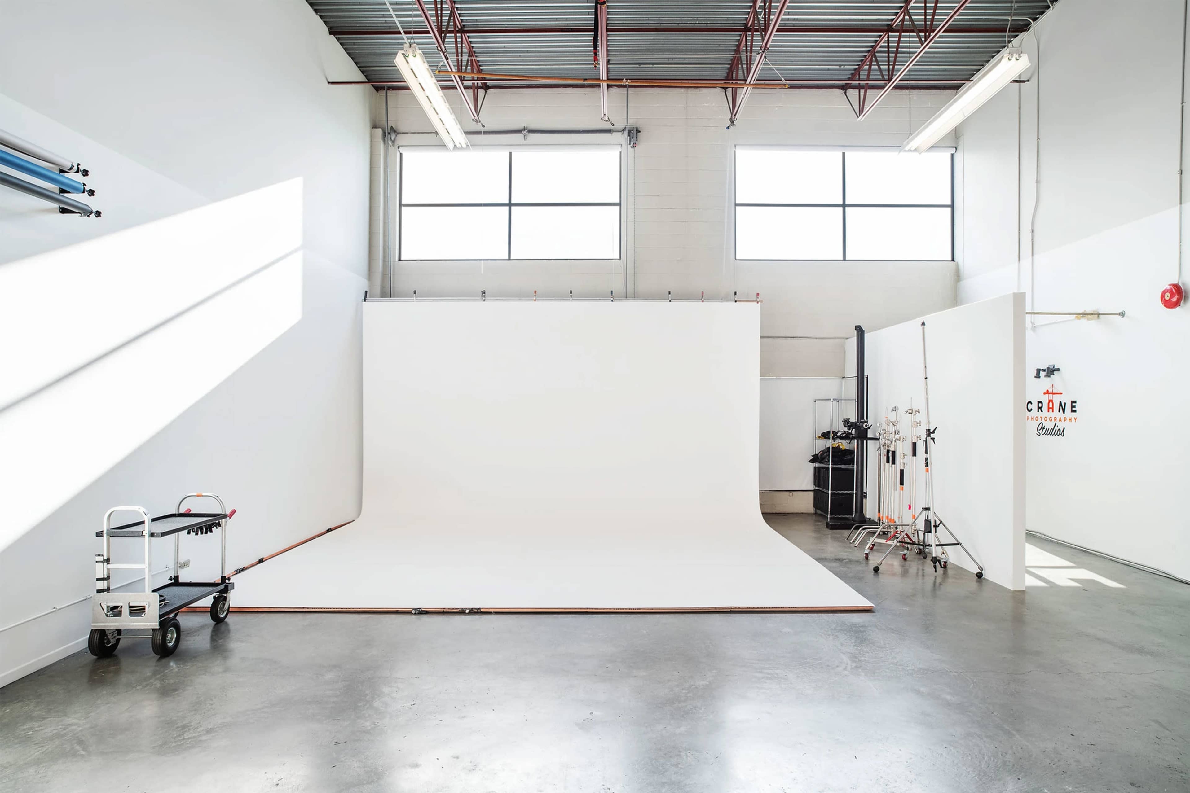 An empty photo studio features a large white backdrop, bright natural light from windows, and gray concrete floors.