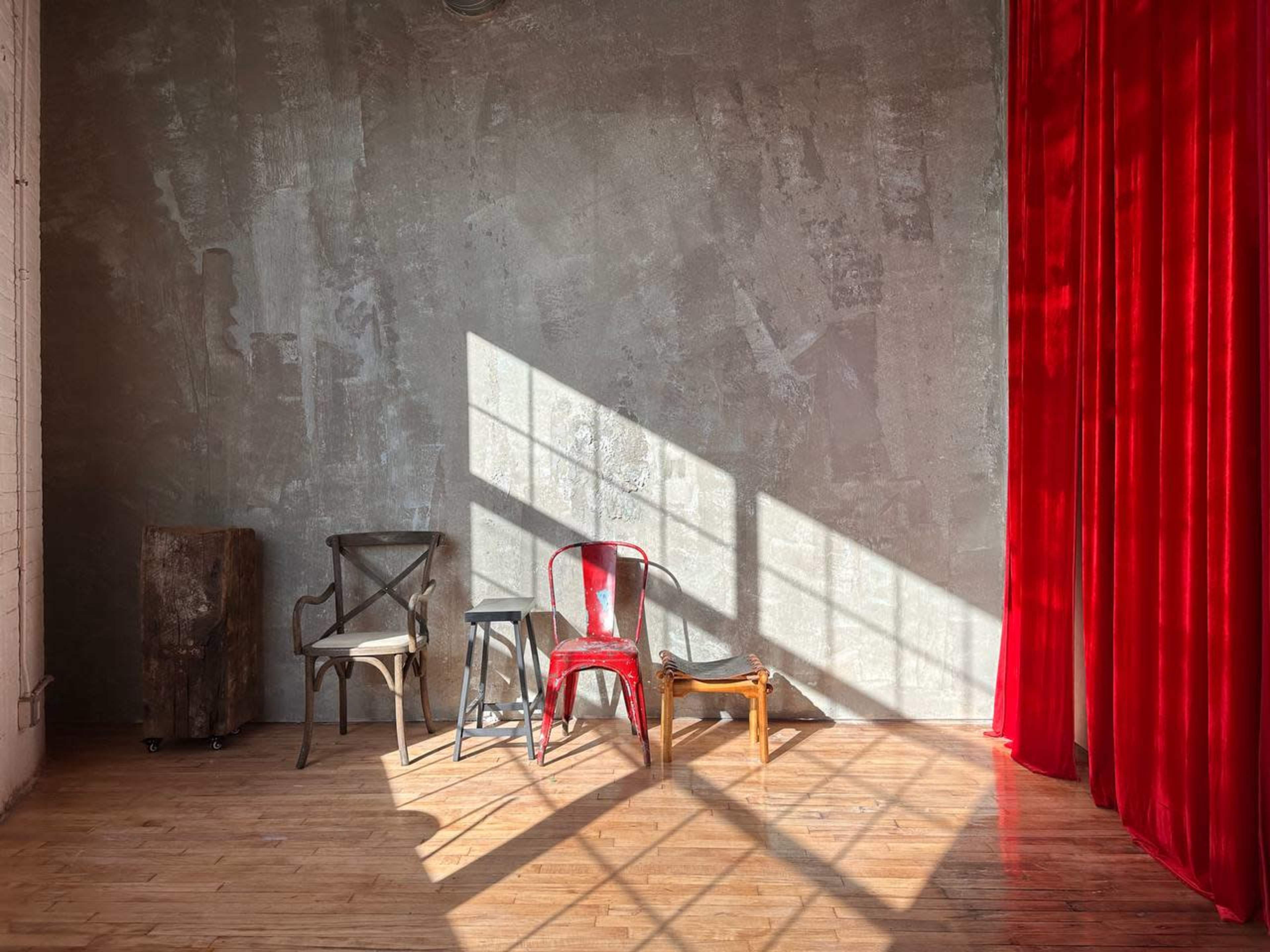 A sunny room with three wooden chairs positioned near a red curtain against a textured wall, illuminated by diagonal sunlight casting shadows on the floor.
