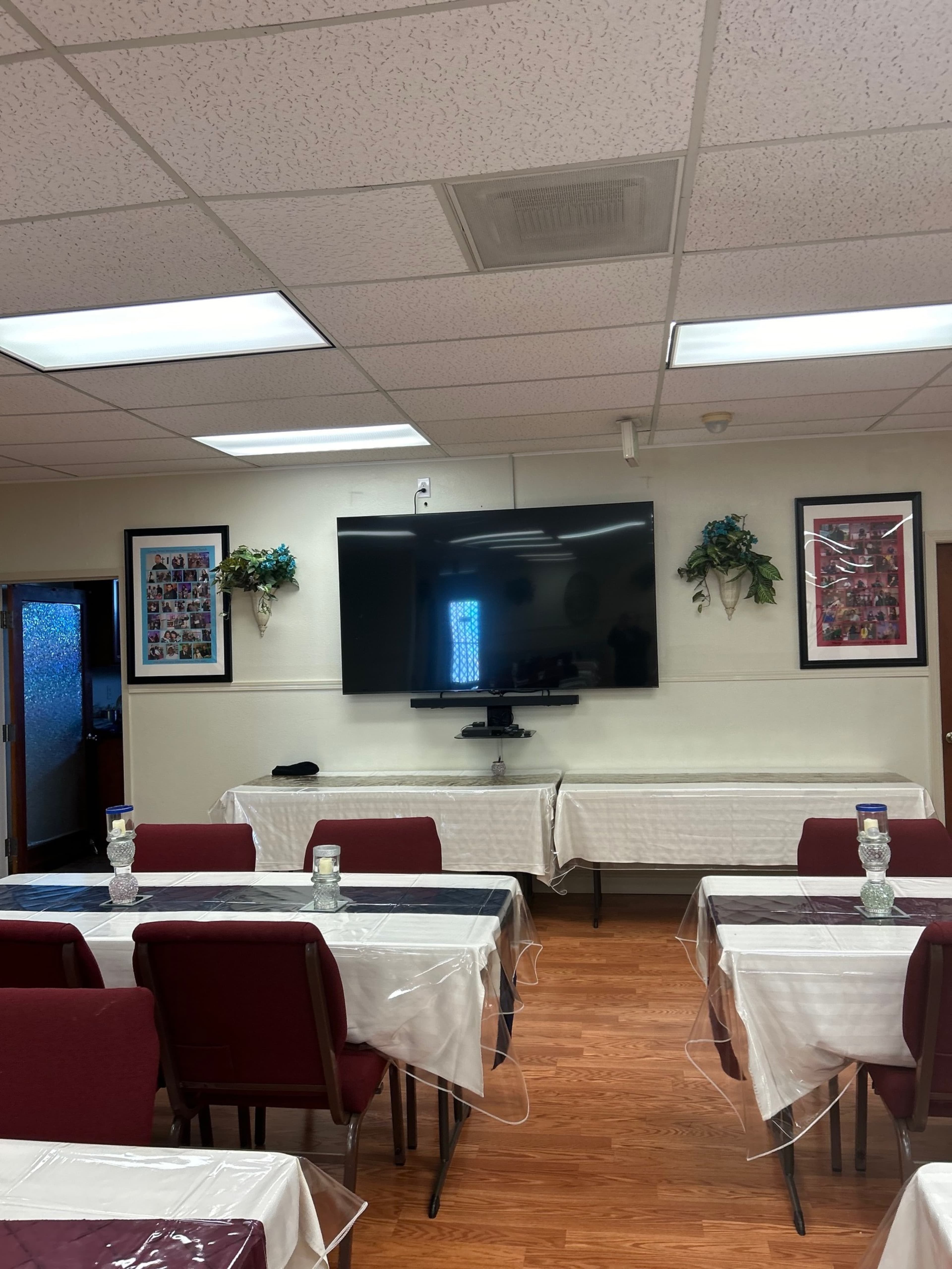 A room set up for an event, featuring tables covered with white tablecloths, red chairs, a large television mounted on the wall, and framed pictures on either side.