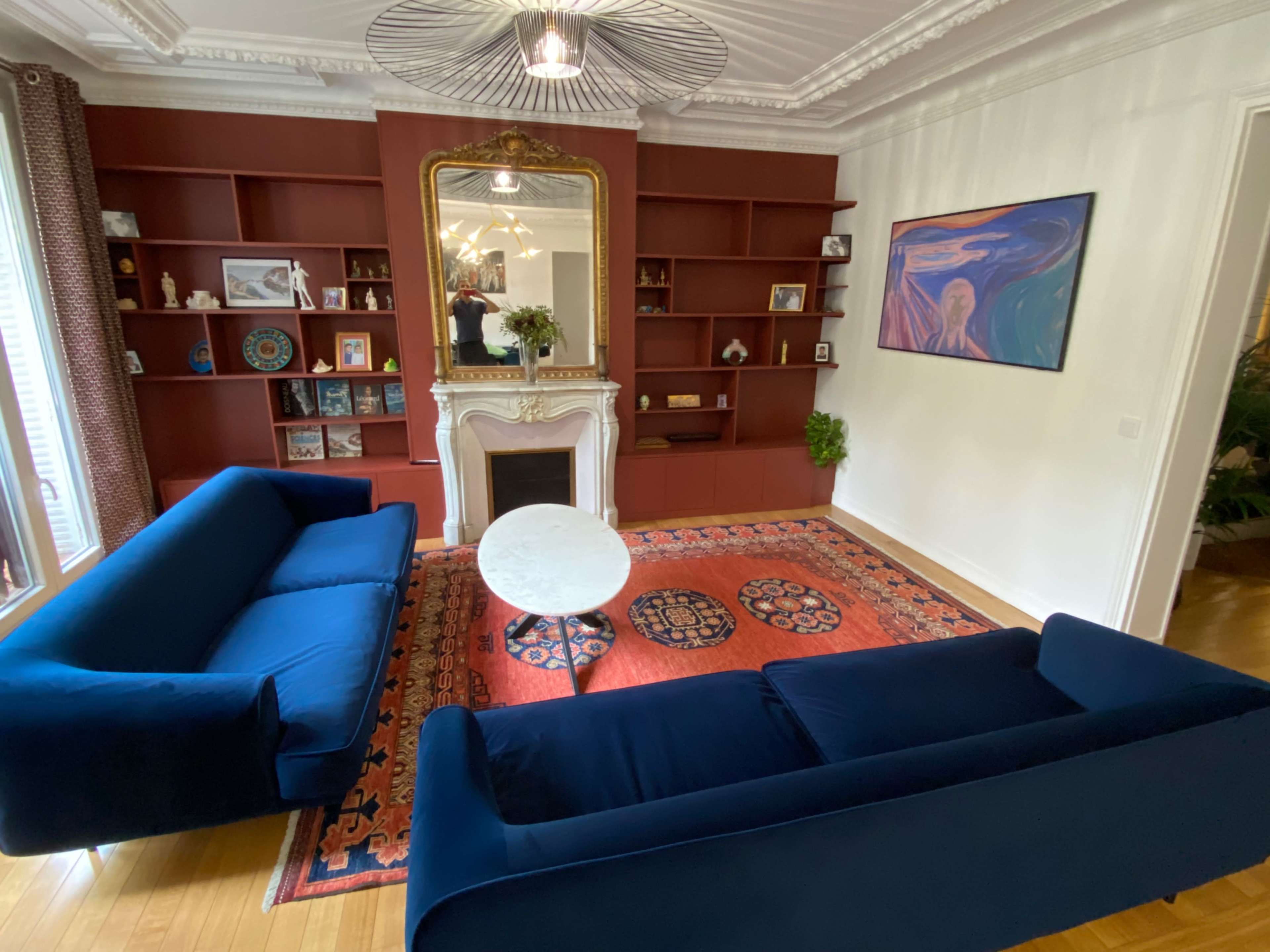 The image shows a cozy living room featuring two blue sofas facing a round coffee table on a patterned rug, with a red accent wall and a mirror above a fireplace.