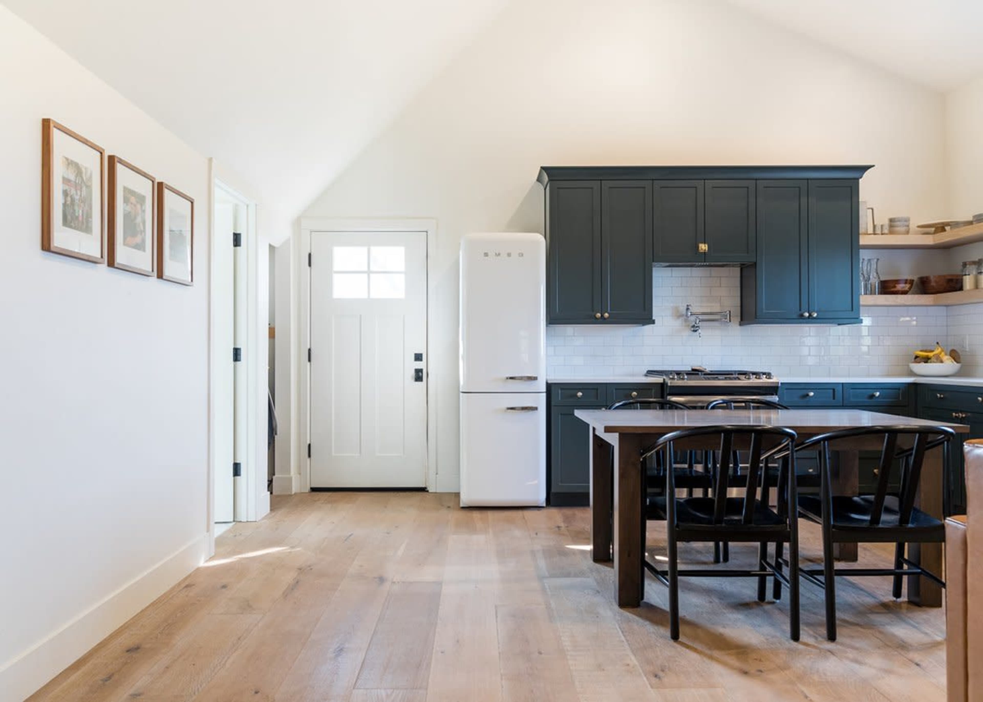 A modern kitchen features a dark blue cabinetry, a white refrigerator, and a wooden dining table with black chairs.