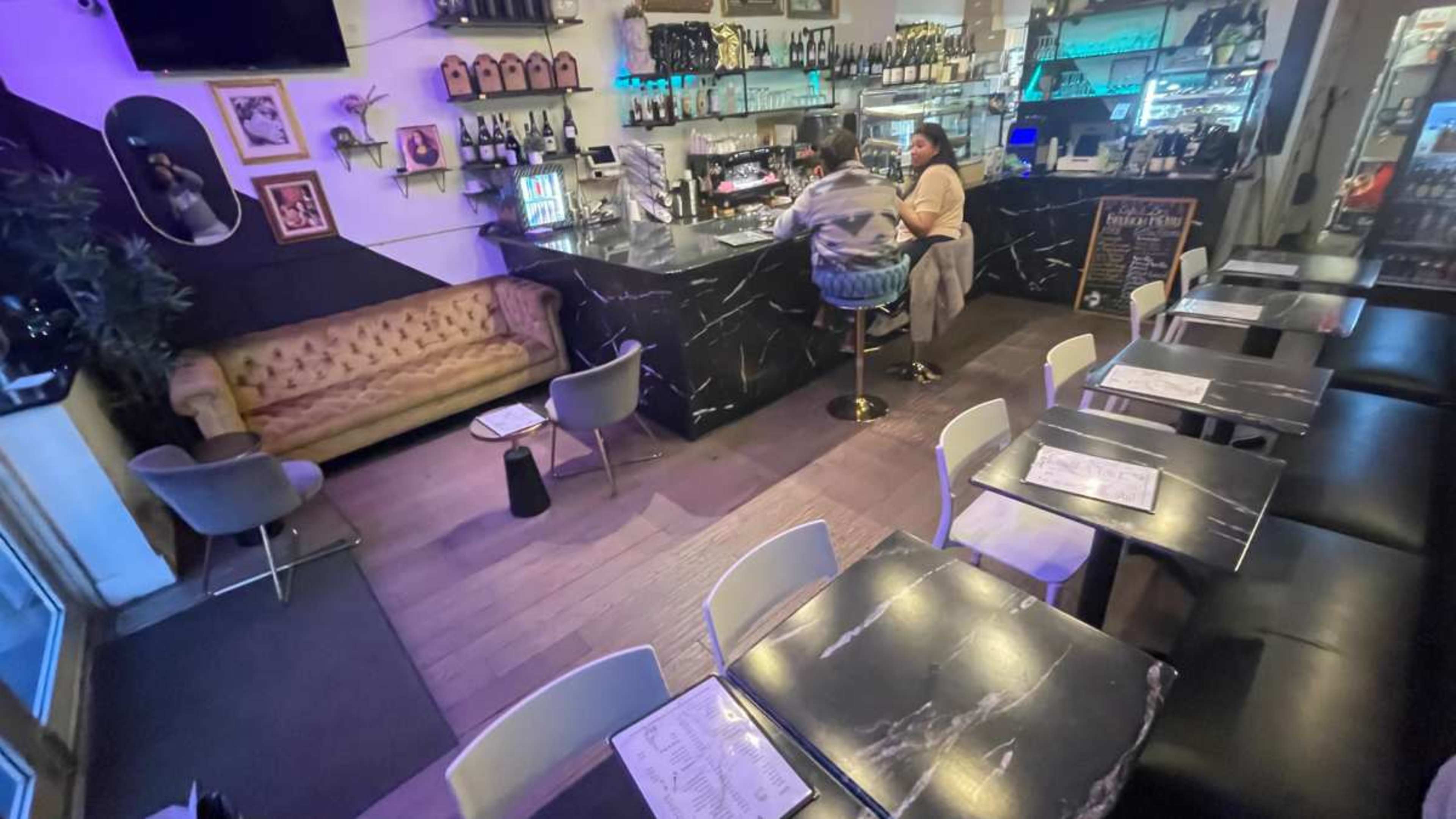 The image shows a bar area with patrons seated at a counter, surrounded by tables and bottles on display.