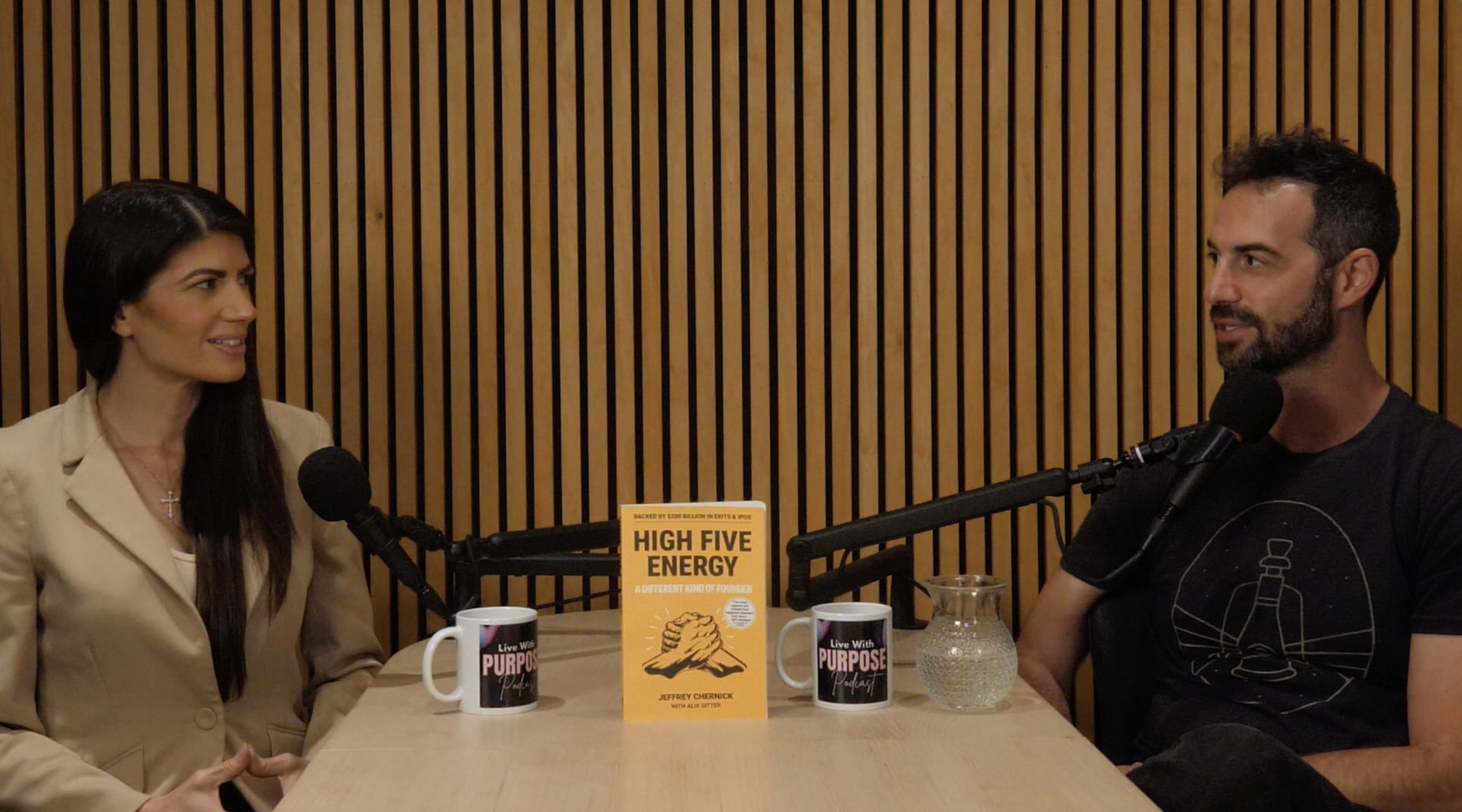 Two people are seated at a table with a book titled "High Five Energy" positioned between them, and both are speaking into microphones in a studio setting with wooden paneling.