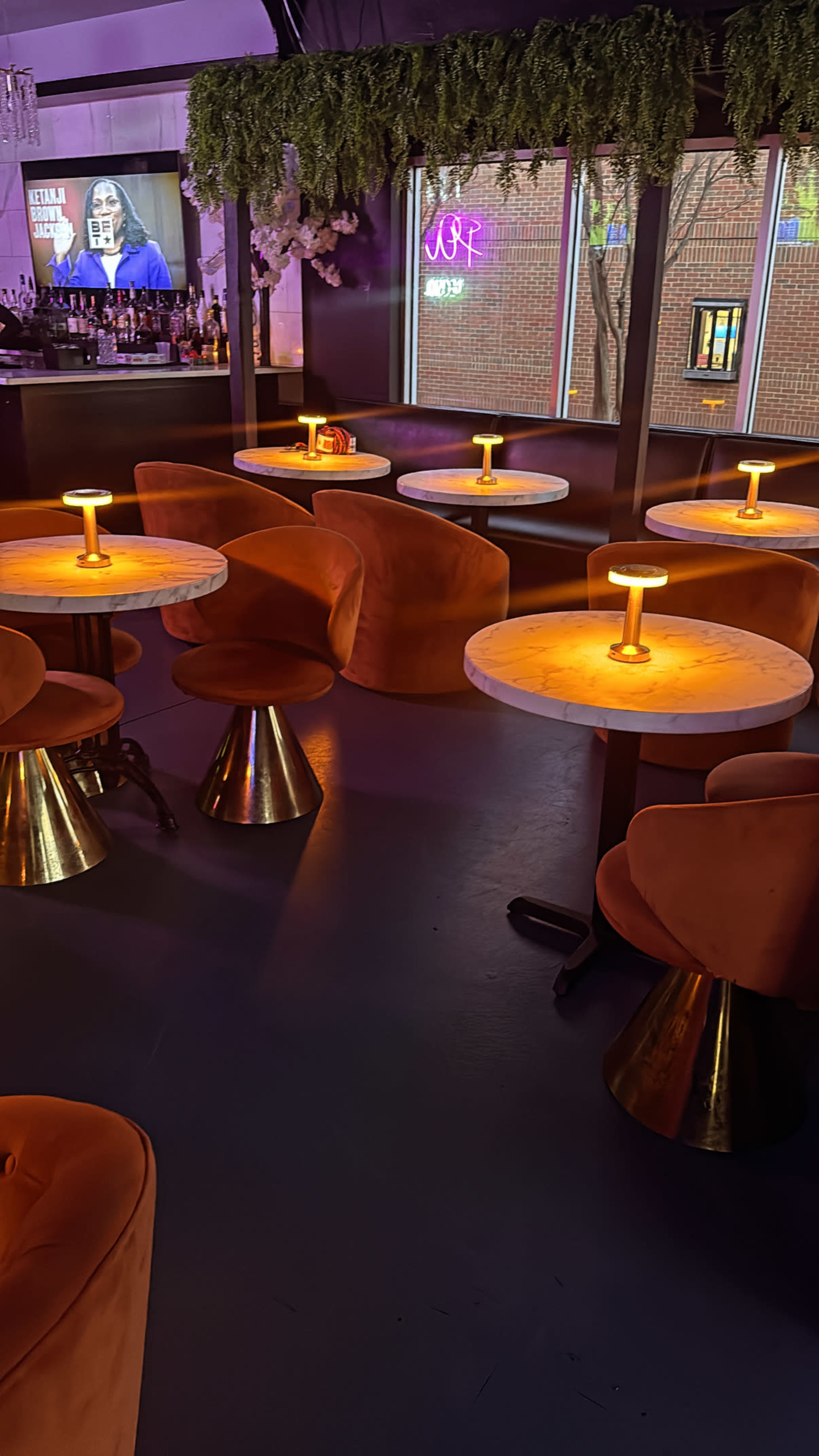 The image shows an indoor bar area with several circular tables topped with glowing lamps and surrounded by plush orange seating.