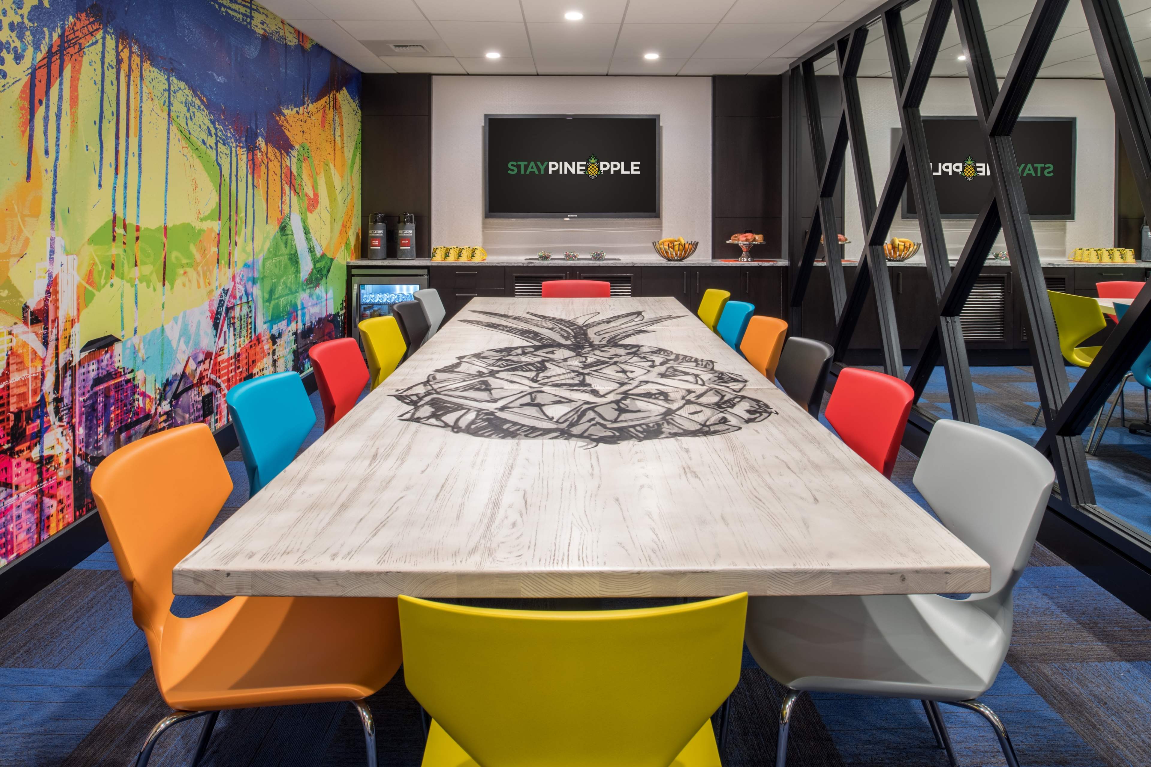 A colorful meeting room featuring a large, artistic table with a pineapple design, surrounded by various brightly colored chairs and a television mounted on the wall.