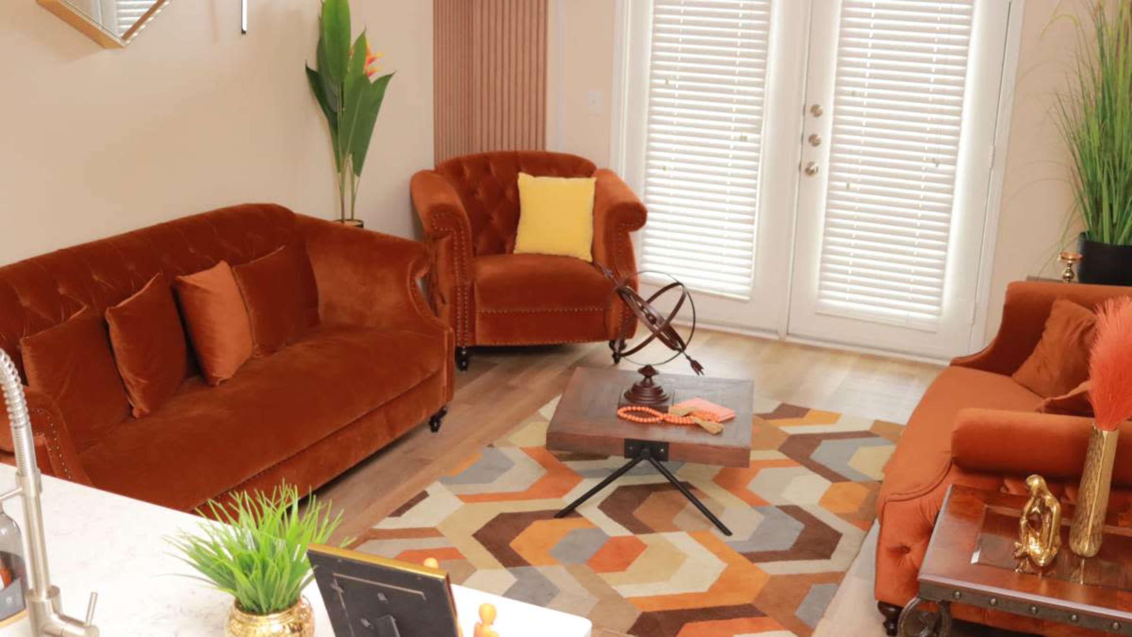 The image shows a living room featuring orange sofas, a geometric patterned rug, and large glass doors opening to an outdoor space.