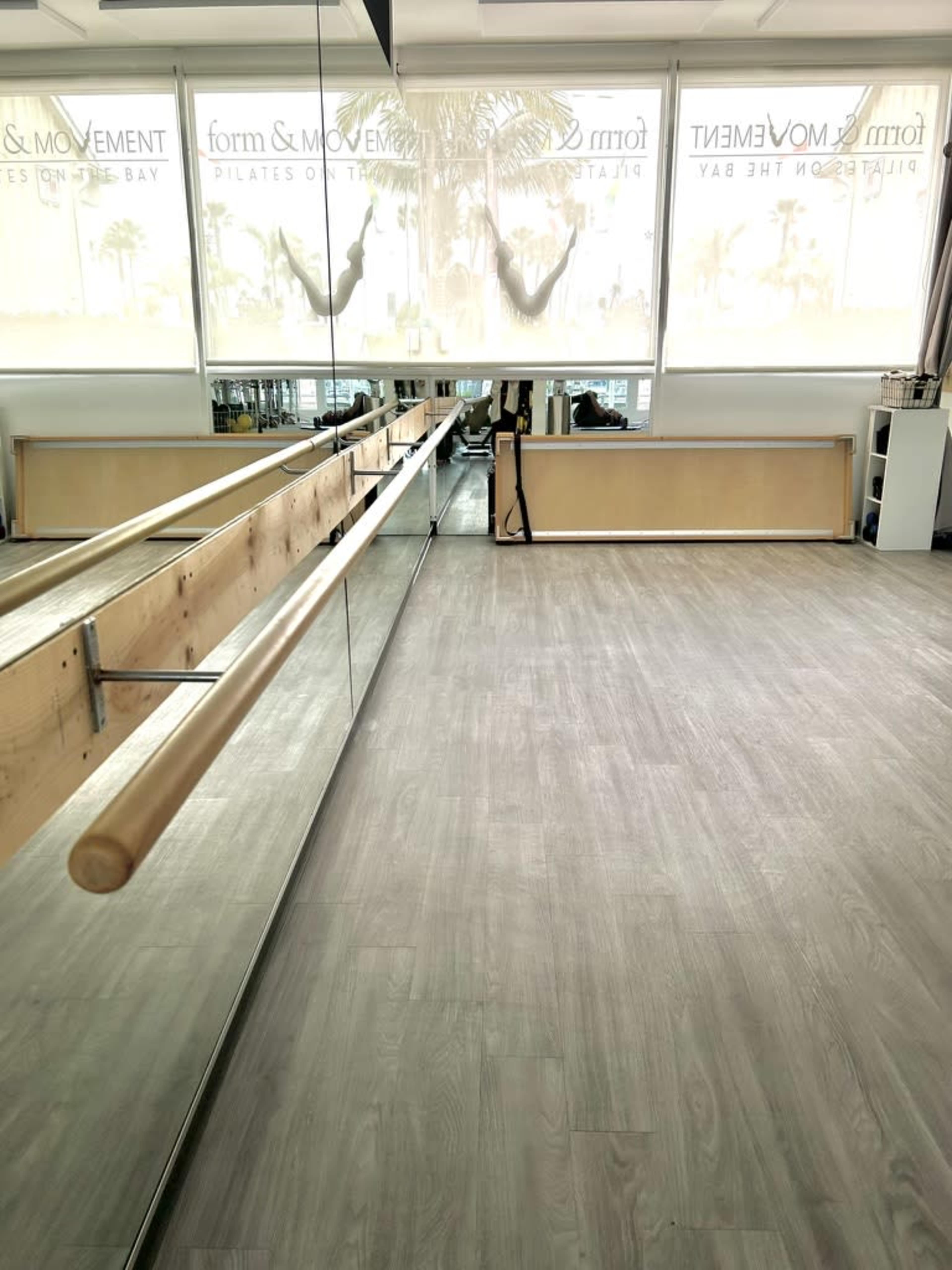 The image shows a Pilates studio with a wooden barre alongside a mirror and large windows allowing natural light to enter.