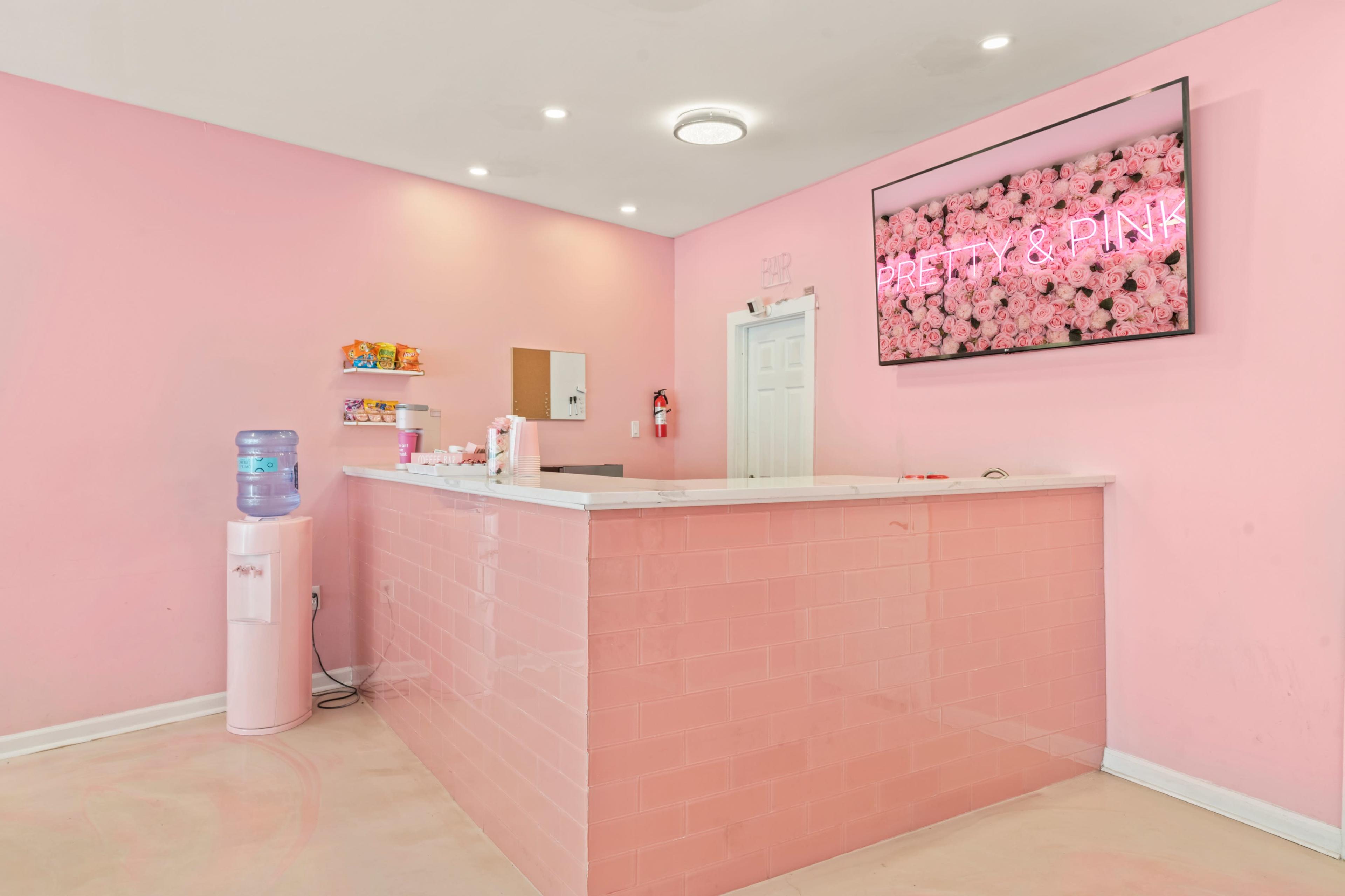A reception area with a pink color scheme, featuring a front desk, a water cooler, and a large screen displaying the phrase "Pretty & Pink" against a backdrop of flowers.