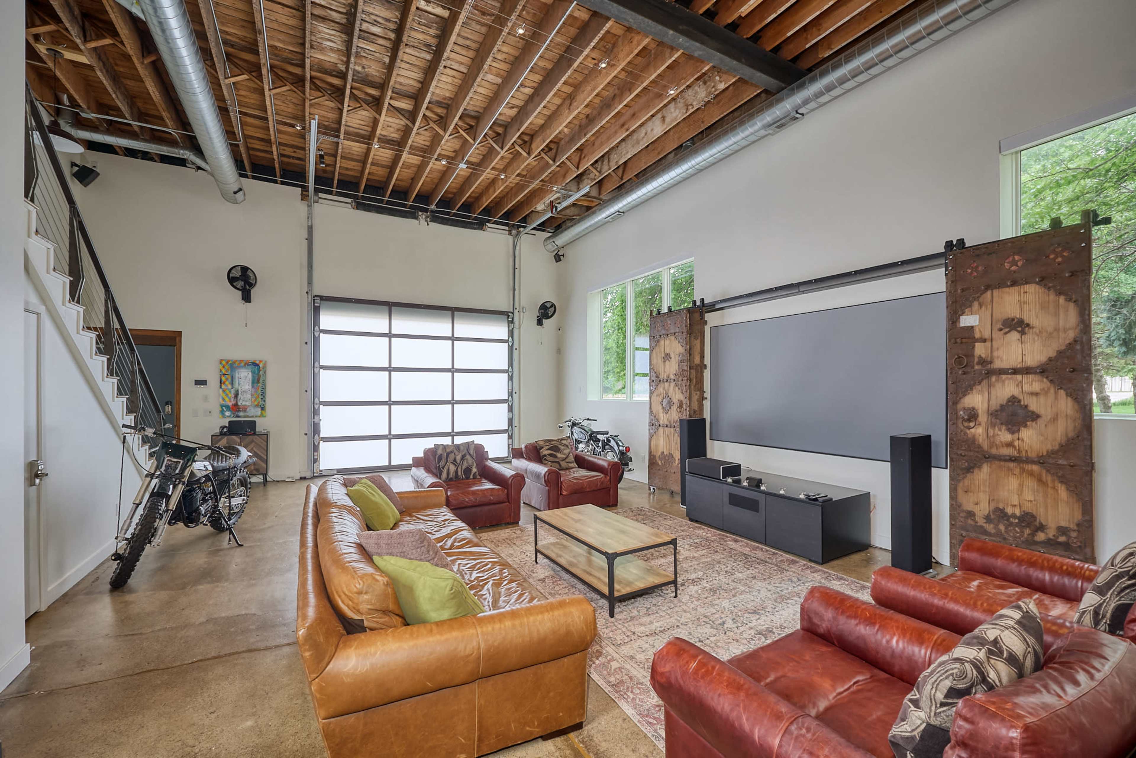 A spacious living area with leather sofas, a coffee table, a large screen on one wall, and a garage door that opens to the outside.